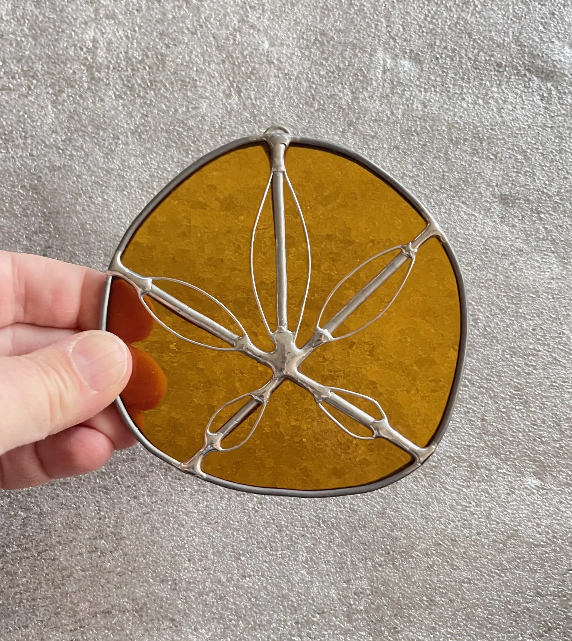 A hand displays the Amber Handmade Stained Glass Sand Dollar with an amber hue and metal outline. This suncatcher contrasts beautifully against a textured gray background, presenting a modern twist on traditional gifts.