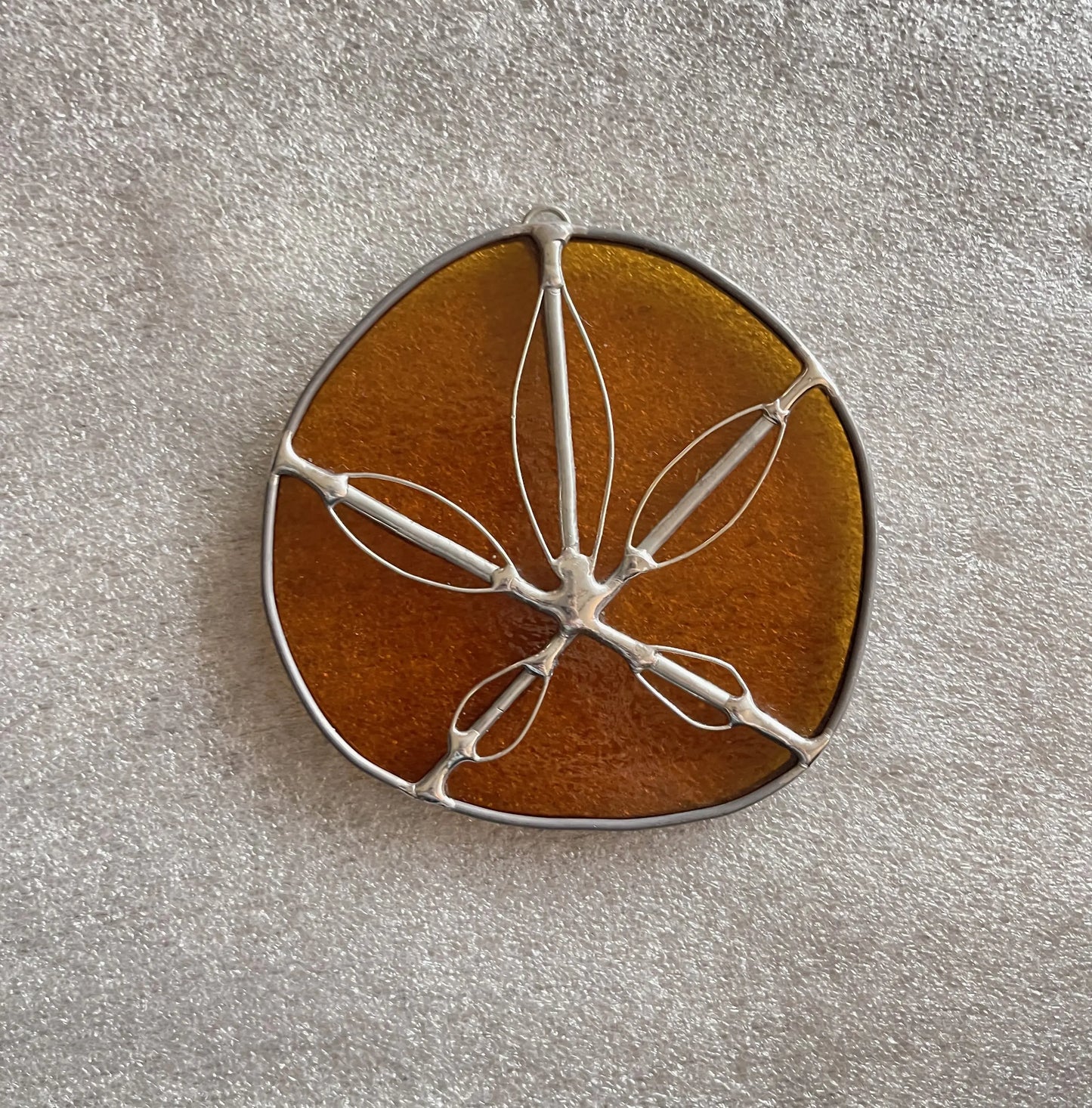 The Amber Handmade Stained Glass Sand Dollar, an oval piece with a silver wire frame and delicate leaf-like patterns, features a translucent amber hue. It rests against a textured light gray surface, offering a unique touch to modern stained glass gifts.