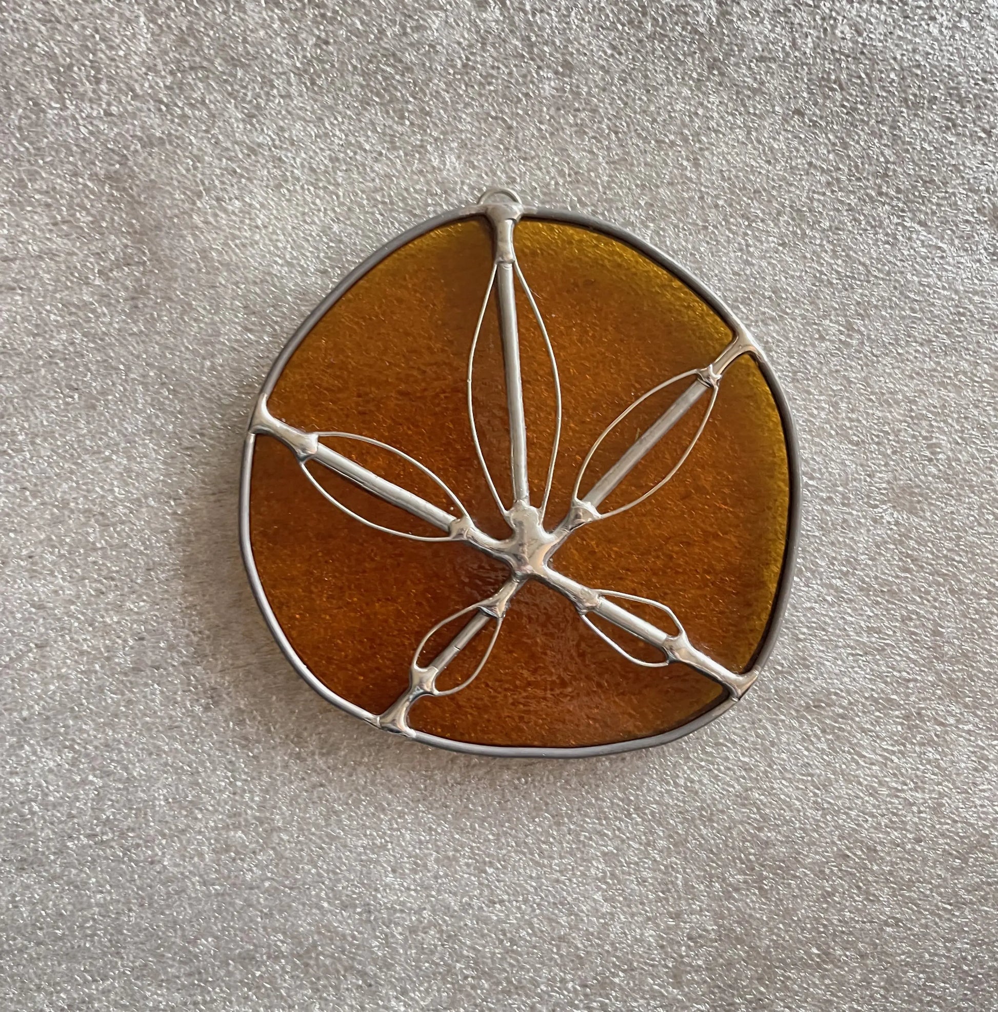 The Amber Handmade Stained Glass Sand Dollar, an oval piece with a silver wire frame and delicate leaf-like patterns, features a translucent amber hue. It rests against a textured light gray surface, offering a unique touch to modern stained glass gifts.