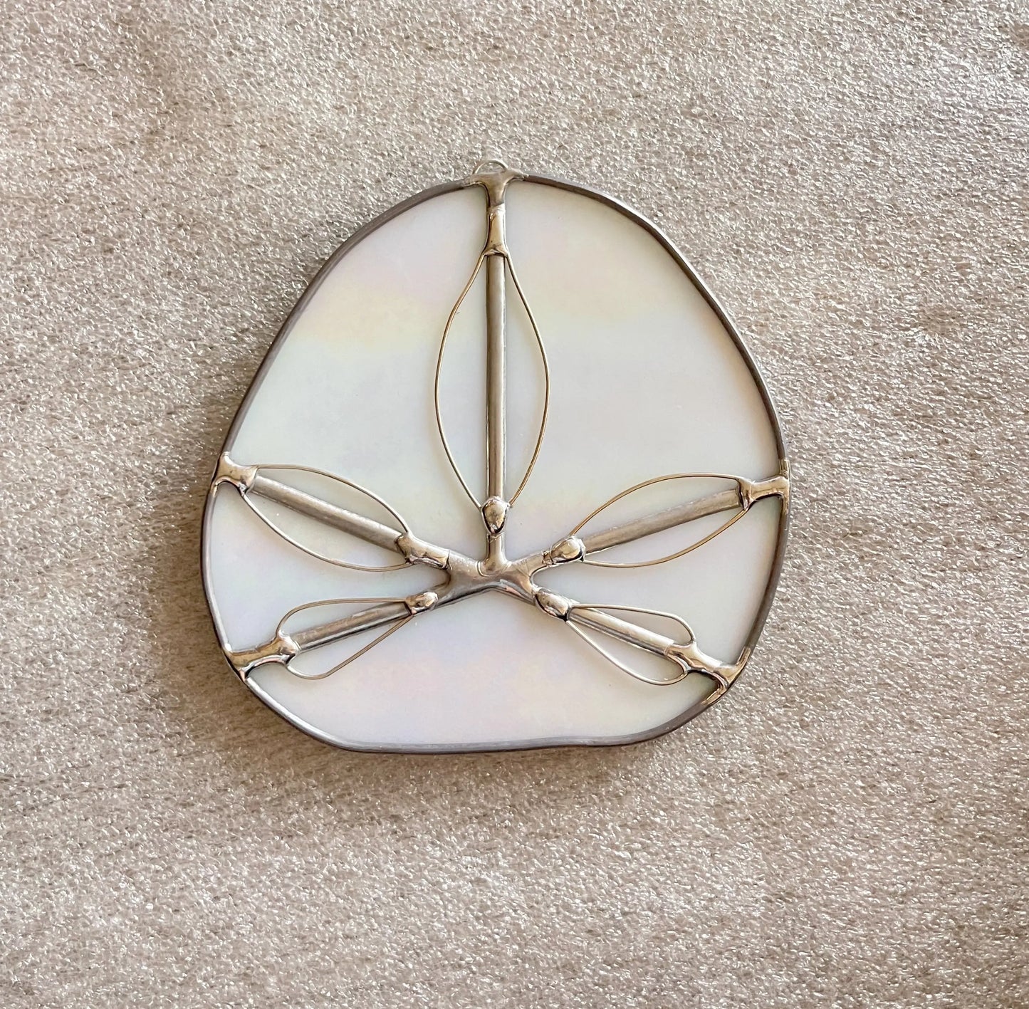 The Iridescent White Sand Dollars feature a delicate, oval mother-of-pearl piece with intricate wire designs on a soft beige textured background. Resembling Pacific Northwest stained glass art, the metal wires form abstract leaf-like shapes evoking handmade stained glass elegance.