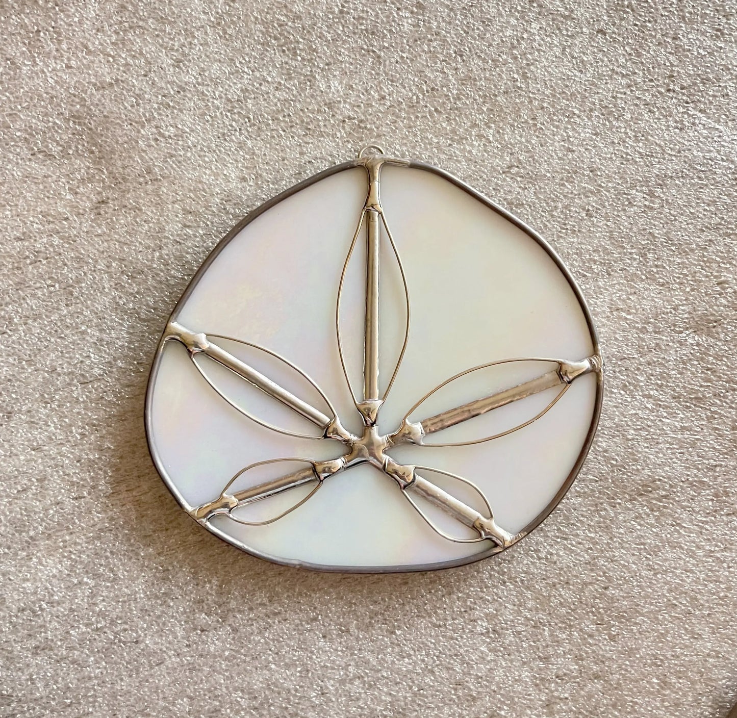 The Iridescent White Sand Dollars handmade stained glass suncatcher features a teardrop shape with a white, iridescent surface. Designed by a Pacific Northwest artist, it boasts abstract interconnected silver-outlined ovals on a textured beige background.
