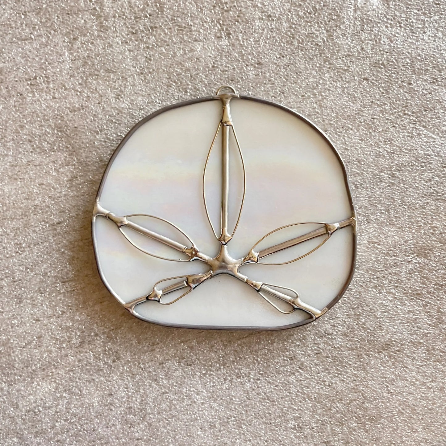 The Iridescent White Sand Dollars is an oval-shaped stained glass piece with metal outlines, crafted by a Pacific Northwest artist. Resembling a floral or star pattern, it rests elegantly on a textured beige surface.