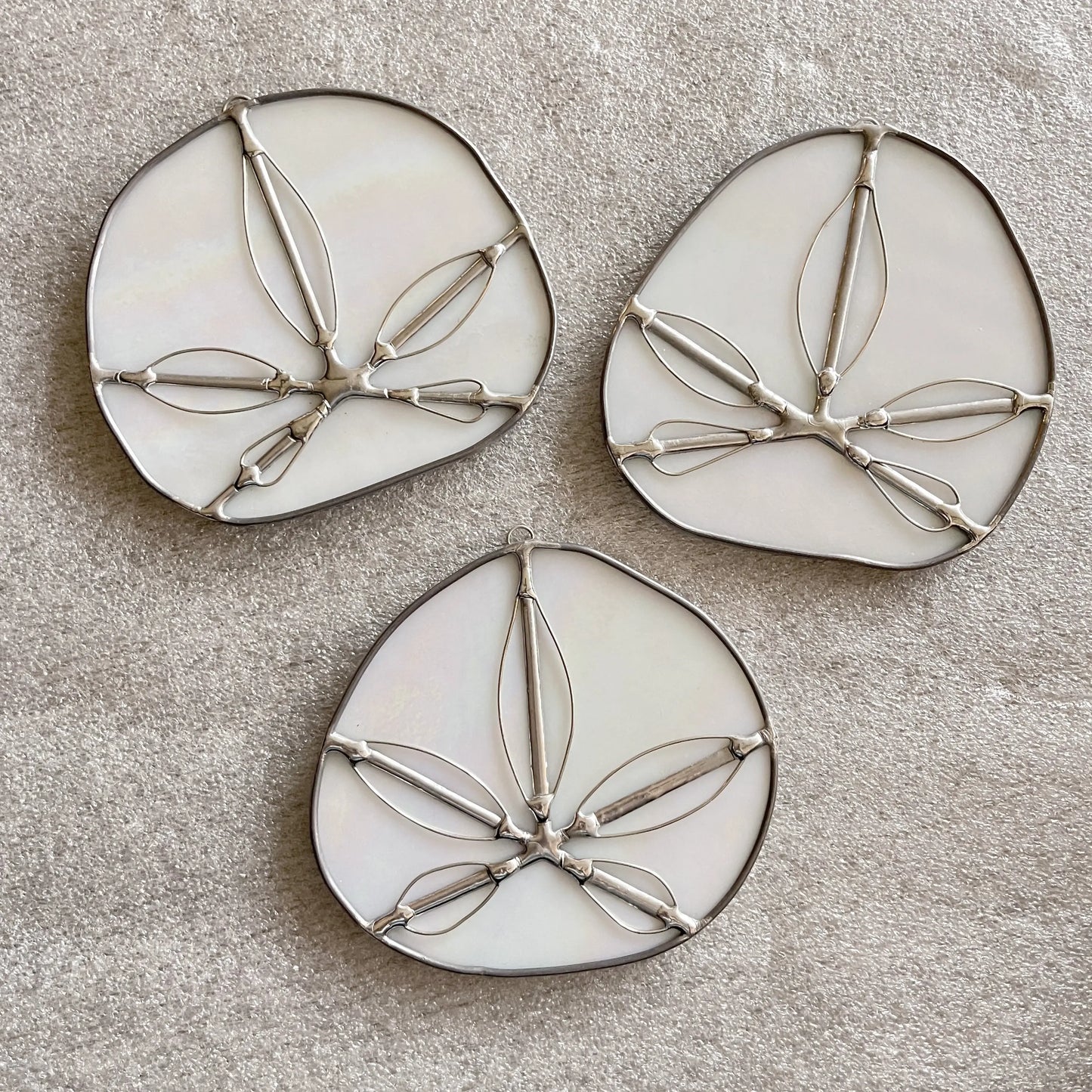 The Iridescent White Sand Dollars collection features three exquisite, handmade stained glass pieces by a Pacific Northwest artist. These decorative suncatchers showcase abstract leaf designs in white and silver, arranged beautifully on a beige textured fabric background.