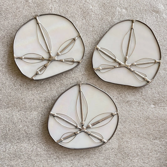 The Iridescent White Sand Dollars collection features three exquisite, handmade stained glass pieces by a Pacific Northwest artist. These decorative suncatchers showcase abstract leaf designs in white and silver, arranged beautifully on a beige textured fabric background.