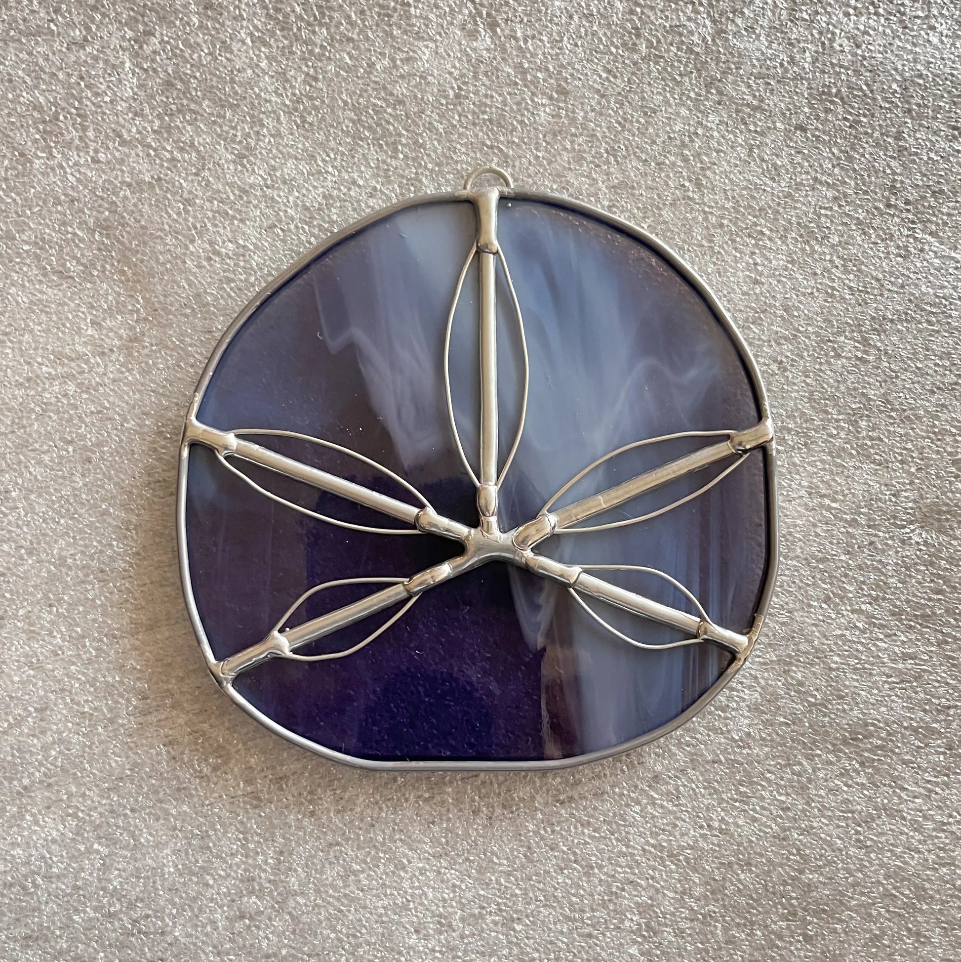 The Purple Streaky Handmade Stained Glass Sand Dollar Ornament, crafted with the Tiffany-method, features a round translucent purple glass design with a silver frame and floral pattern. It rests elegantly against a light tan textured surface.