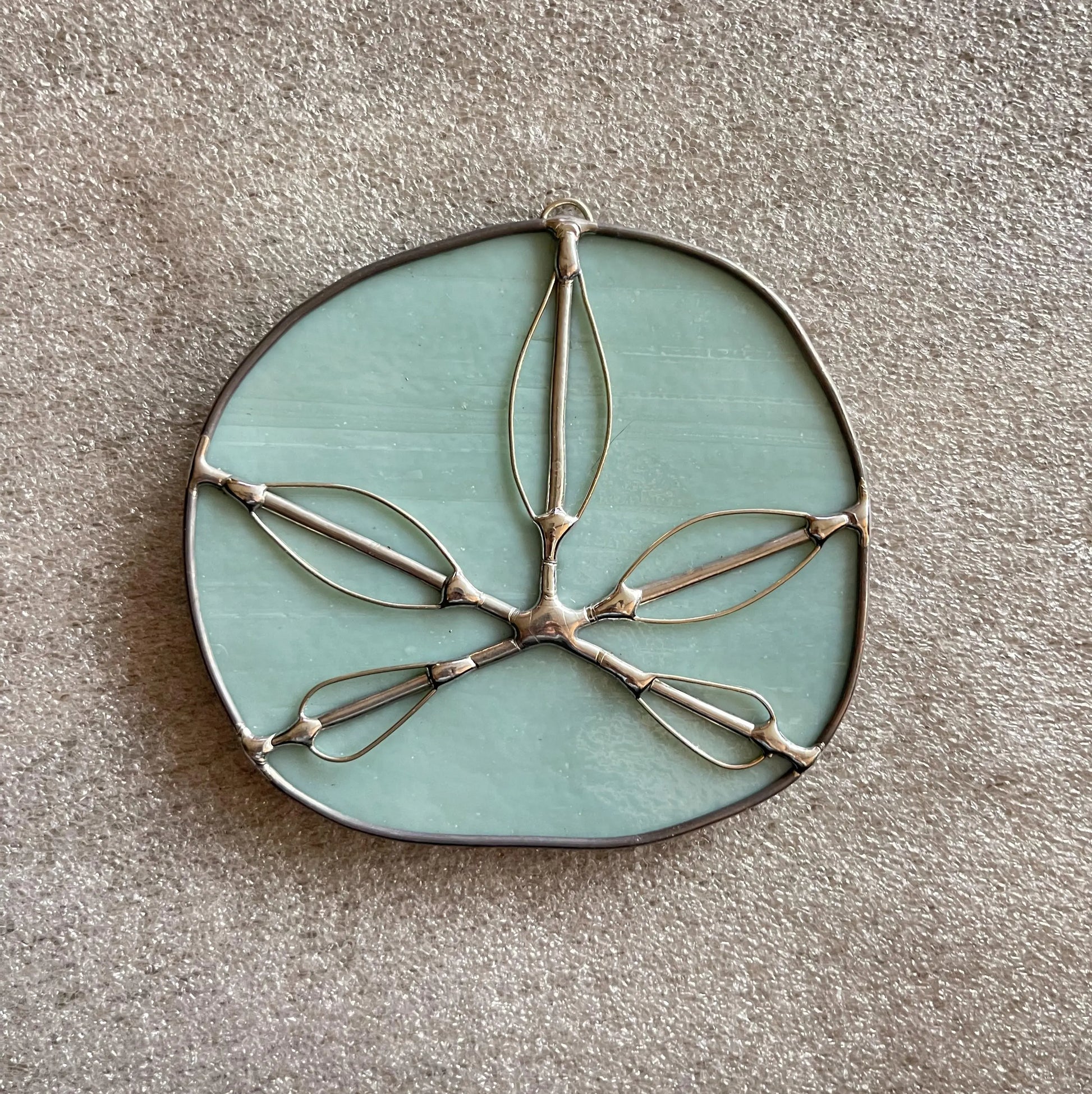 The Turquoise Handmade Stained Glass Sand Dollar is a circular decorative piece featuring a light green, cloudy glass surface encased in metal. It mimics a sand dollar with five thin, elongated metal loops forming a star-like pattern on the front.