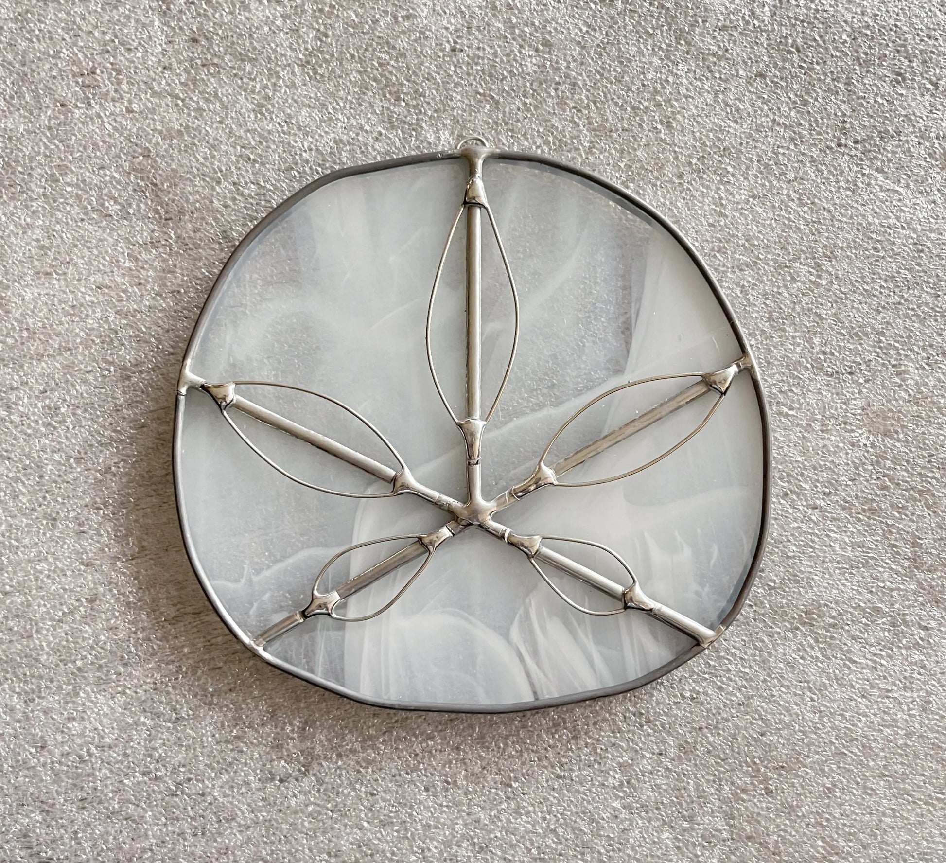 A round, iridescent Translucent White Sand Dollars handmade stained glass ornament with a silver wire leaf design rests on textured light fabric. Crafted using the Tiffany-method, its pale hues add an ethereal touch to this exquisite gift.