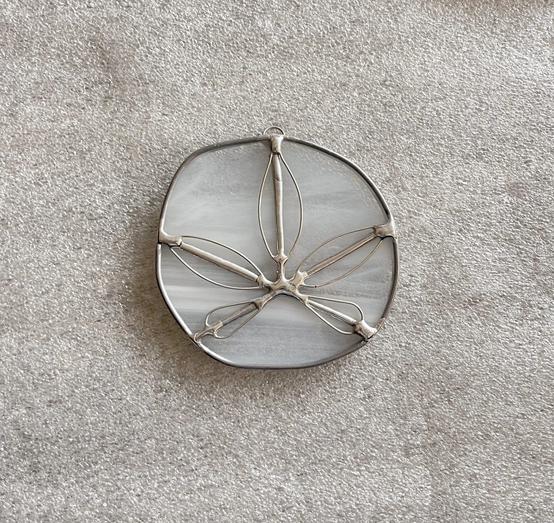 A Translucent White Sand Dollars handmade stained glass ornament with silver outlines, designed in an abstract flower shape, rests elegantly on a textured, light-colored surface.
