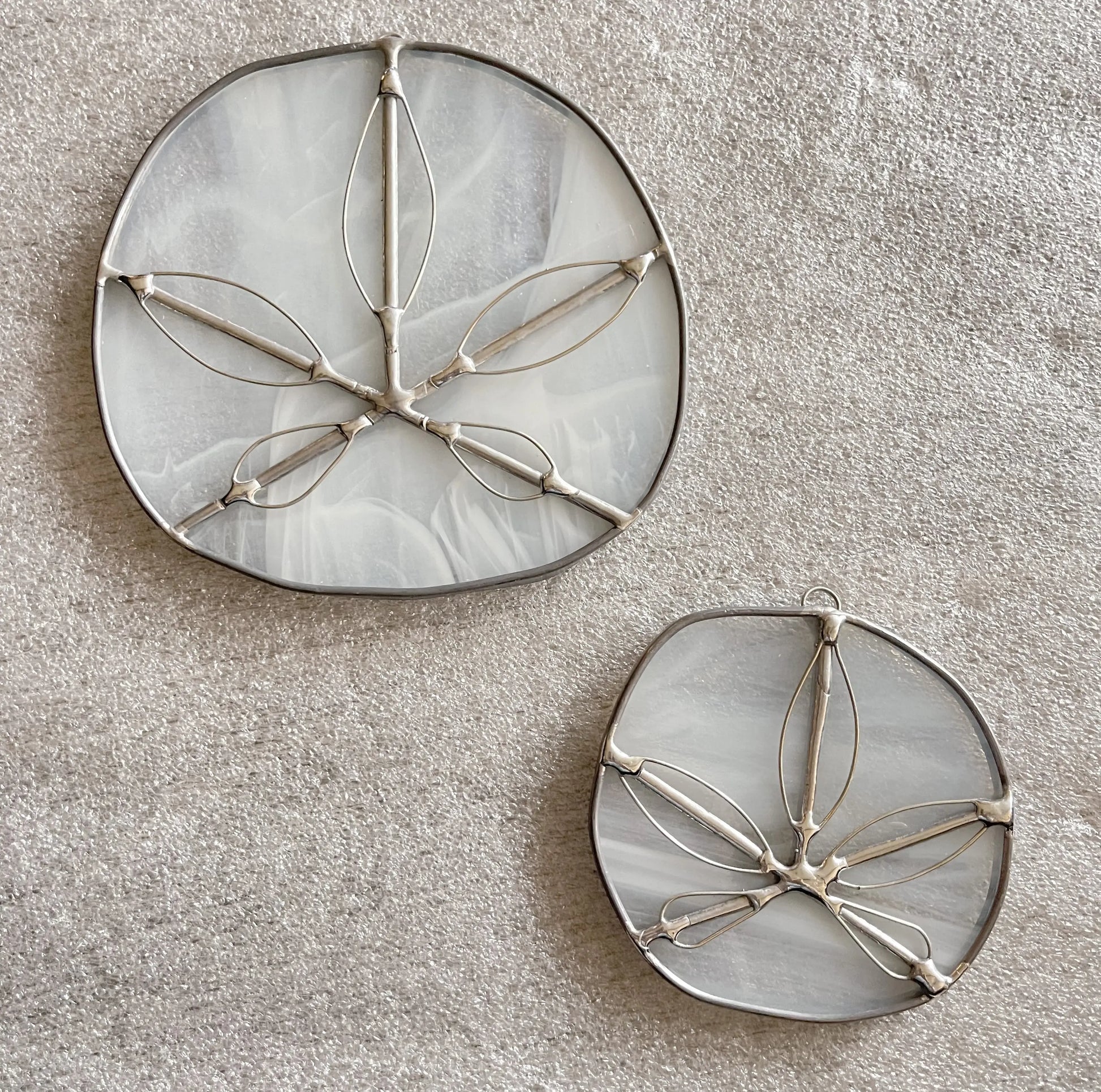 Two round Translucent White Sand Dollars handmade suncatchers, crafted using the Tiffany-method, rest on a textured light brown surface. Each features a leaf pattern with metal outlines on a cloudy background. The set includes one larger ornament beside its smaller counterpart.