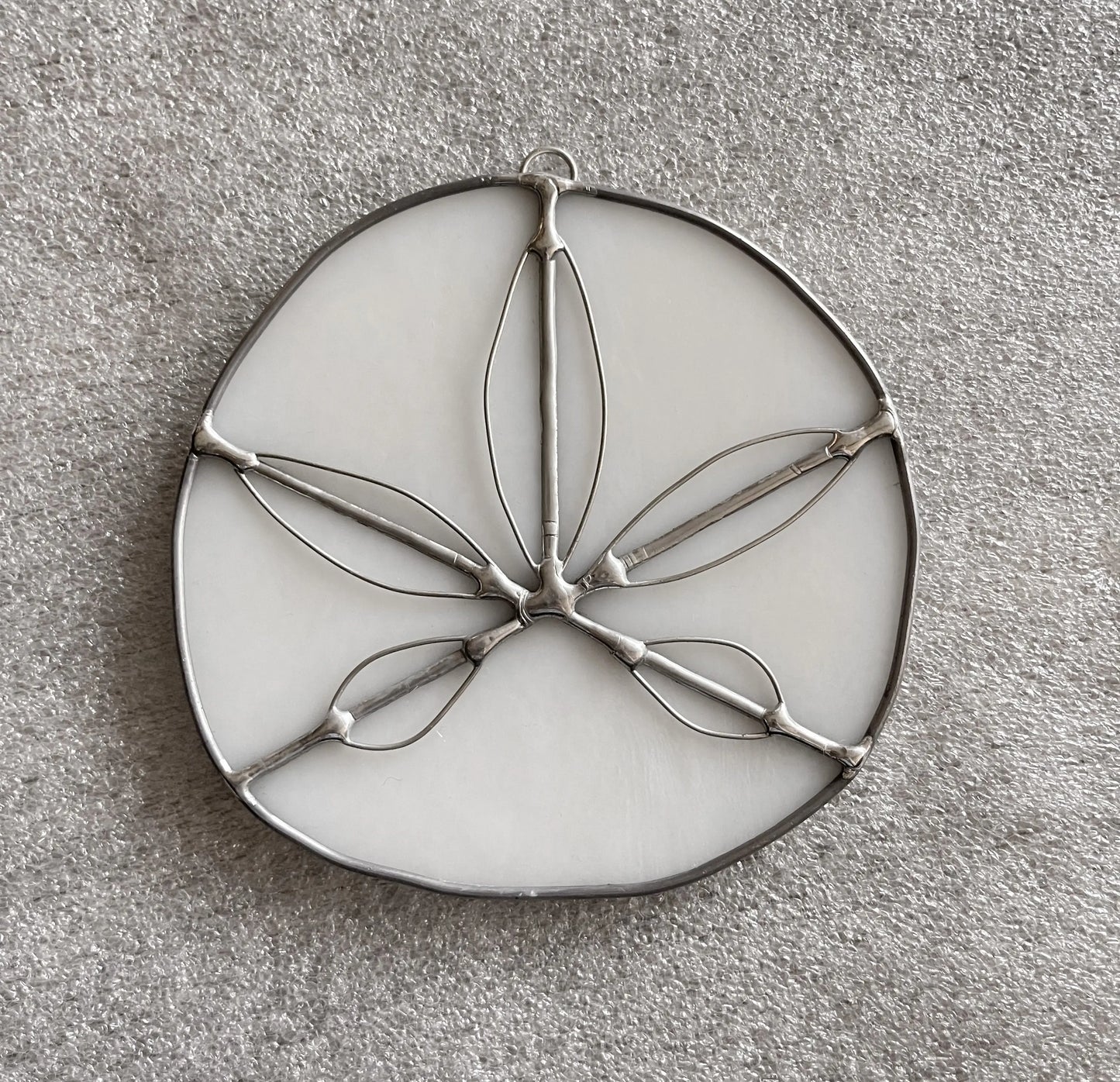 The Handmade Stained Glass Sand Dollars - White is a round, handmade piece by a Pacific Northwest artist featuring a white background with floral or leaf metal outlines and five elongated petal-like shapes radiating from the center on a gray textured surface.
