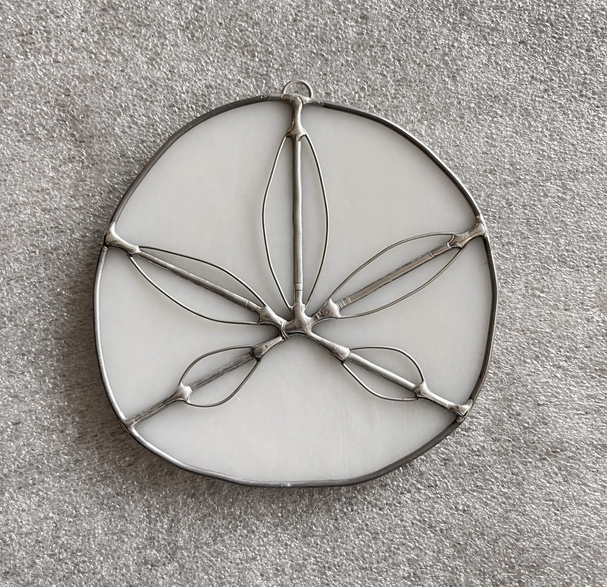The Handmade Stained Glass Sand Dollars - White is a round, handmade piece by a Pacific Northwest artist featuring a white background with floral or leaf metal outlines and five elongated petal-like shapes radiating from the center on a gray textured surface.
