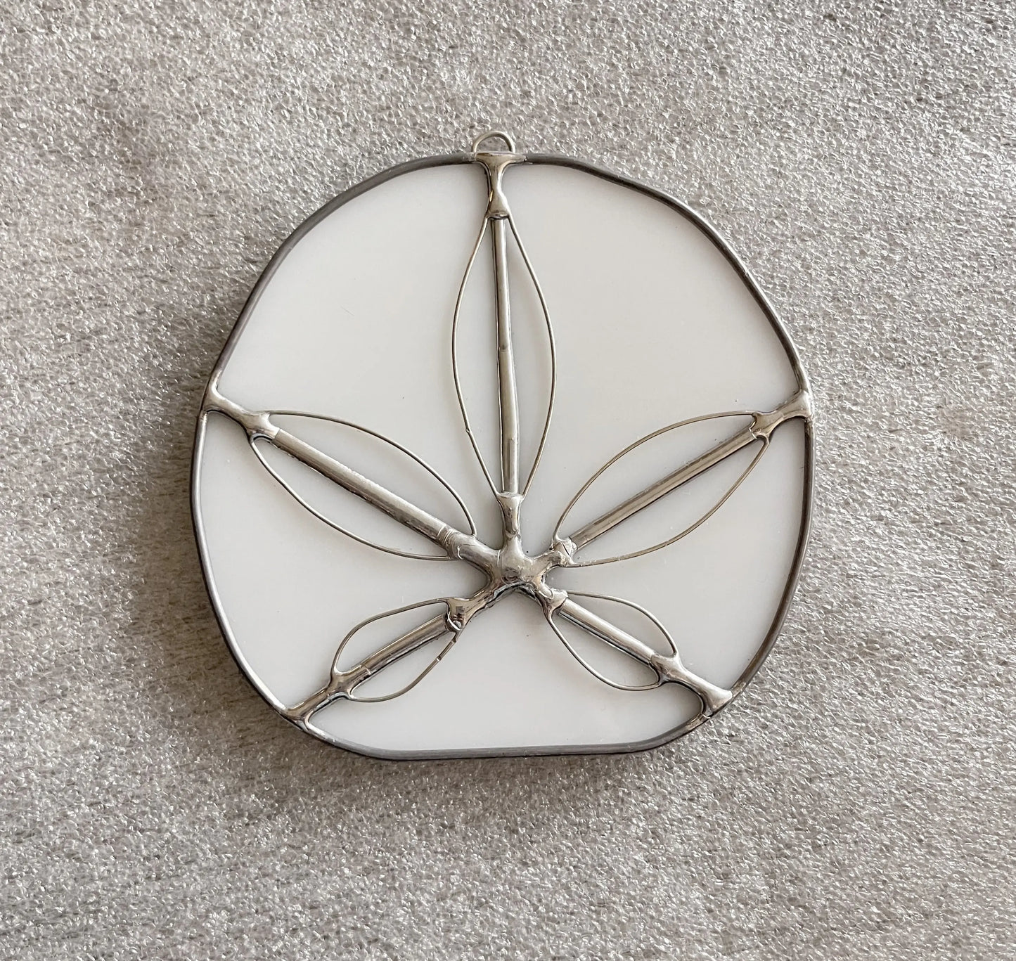 The Handmade Stained Glass Sand Dollars - White features an oval base like a sand dollar with metal outlines of leaves or petals set on a textured, light gray surface, showcasing intricate craftsmanship inspired by Pacific Northwest artists.