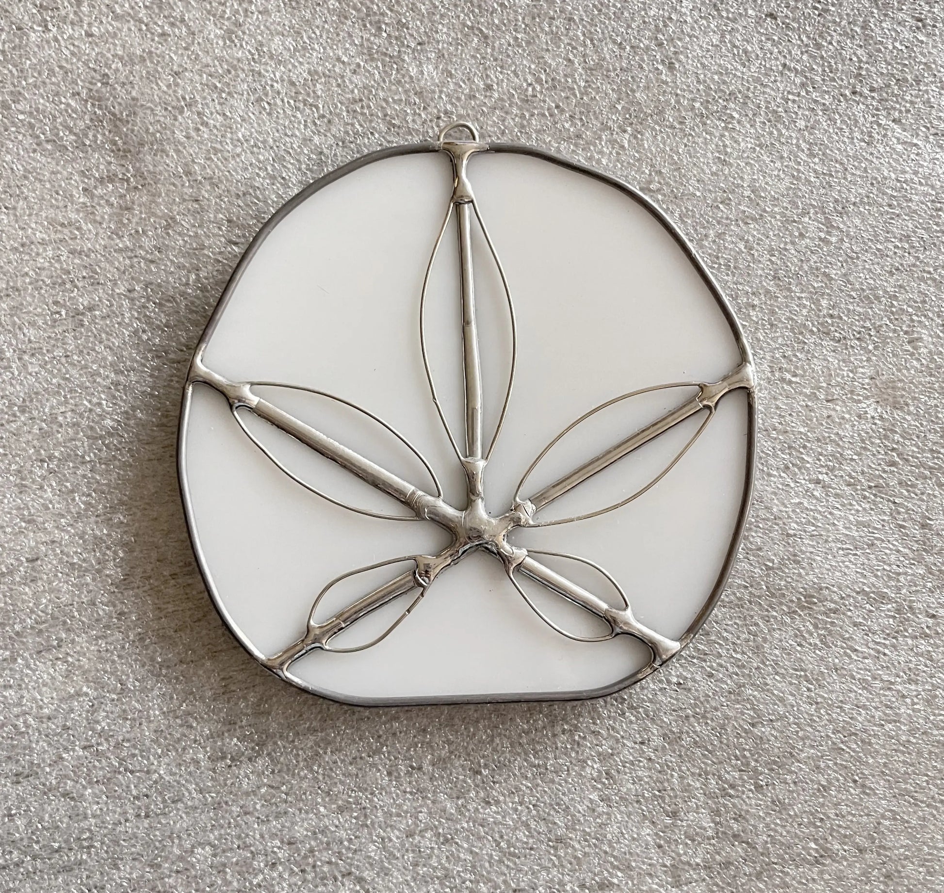 The Handmade Stained Glass Sand Dollars - White features an oval base like a sand dollar with metal outlines of leaves or petals set on a textured, light gray surface, showcasing intricate craftsmanship inspired by Pacific Northwest artists.