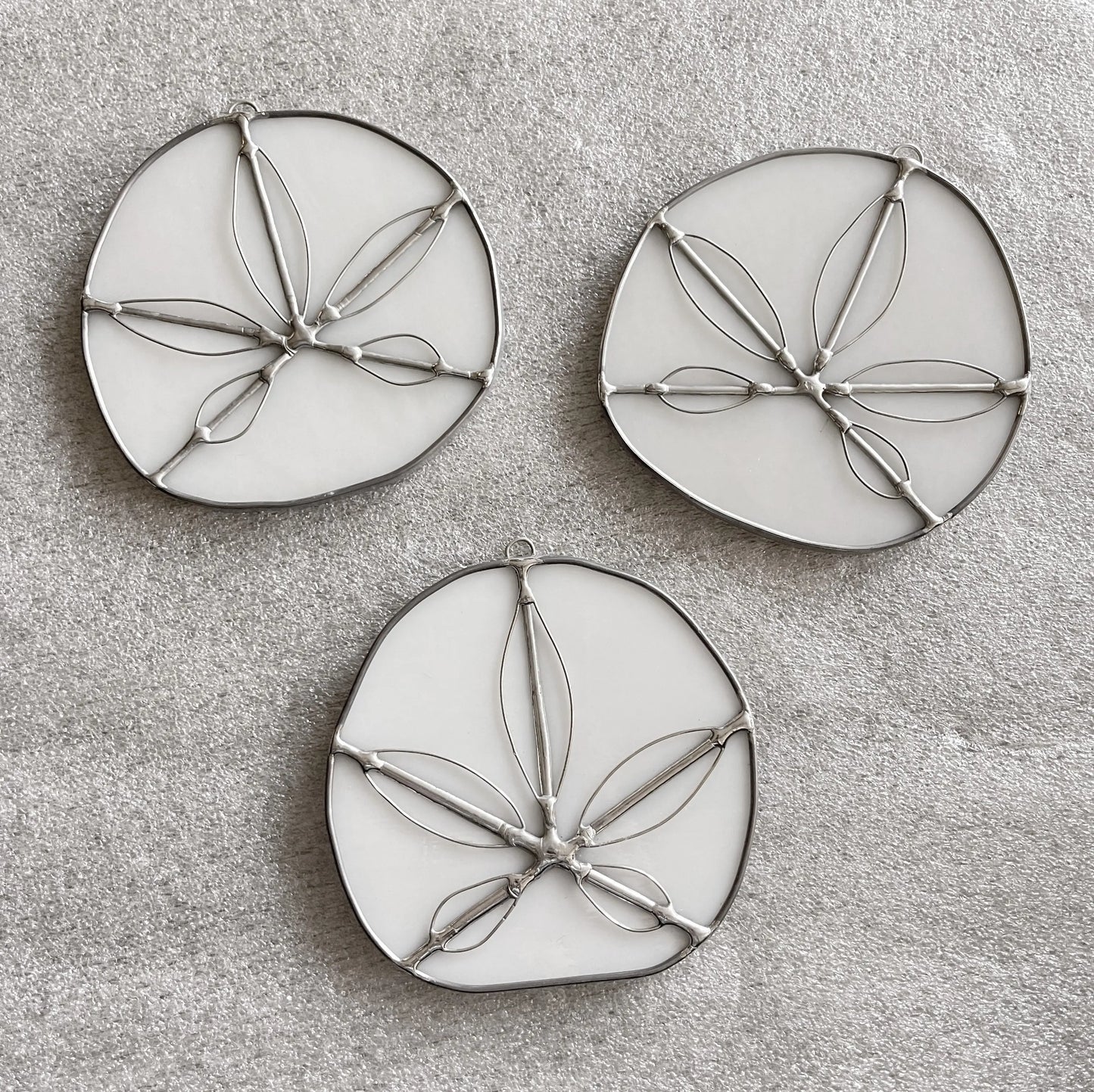 The Handmade Stained Glass Sand Dollars - White feature three round, white ornaments with metal outlines resembling sand dollars, arranged on a light gray textured surface. Crafted by a Pacific Northwest artist, these unique suncatchers bring coastal charm to any space.