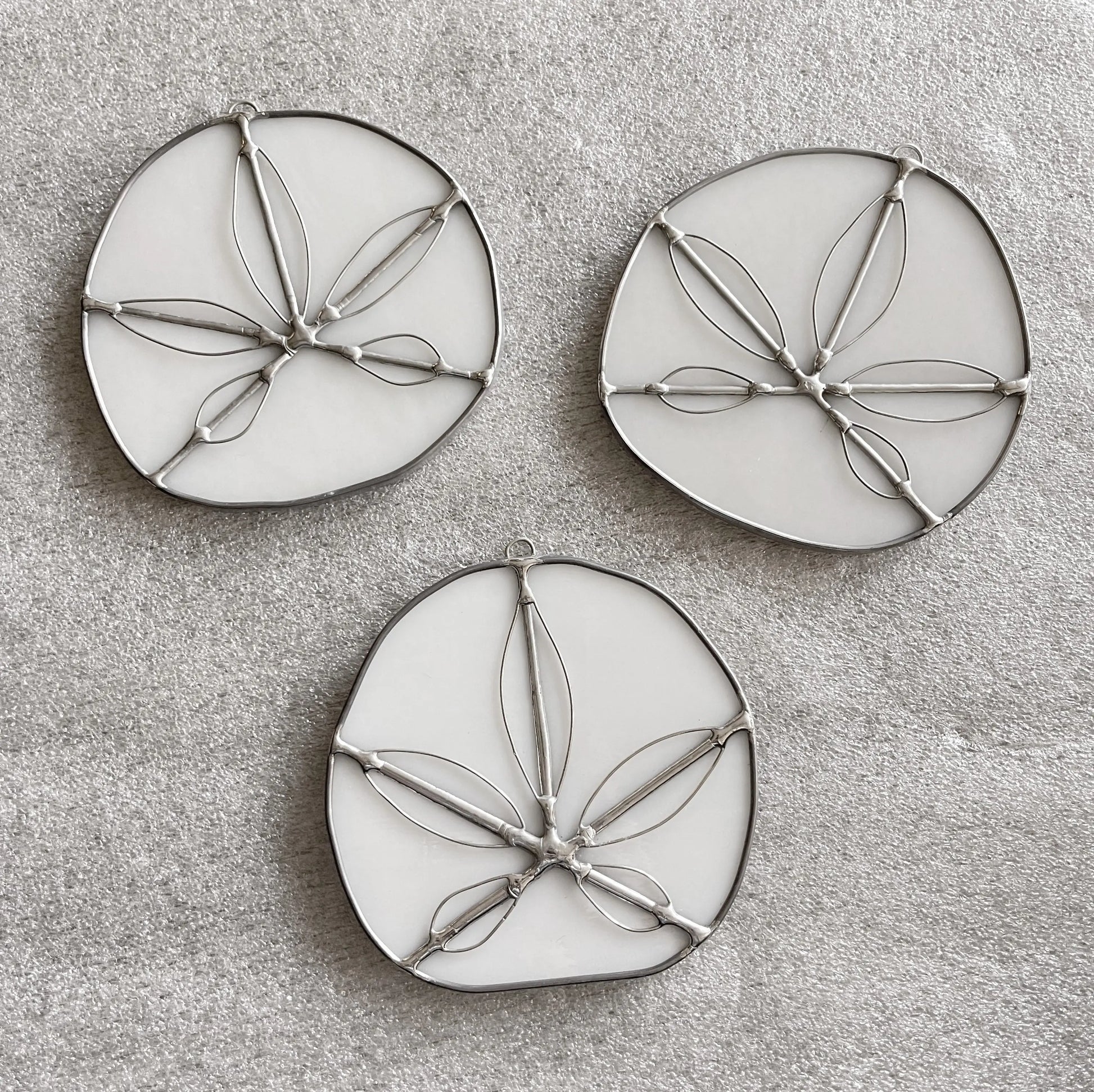 The Handmade Stained Glass Sand Dollars - White feature three round, white ornaments with metal outlines resembling sand dollars, arranged on a light gray textured surface. Crafted by a Pacific Northwest artist, these unique suncatchers bring coastal charm to any space.