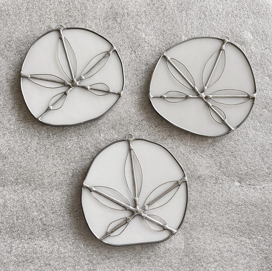 The Handmade Stained Glass Sand Dollars - White feature three round, white ornaments with metal outlines resembling sand dollars, arranged on a light gray textured surface. Crafted by a Pacific Northwest artist, these unique suncatchers bring coastal charm to any space.