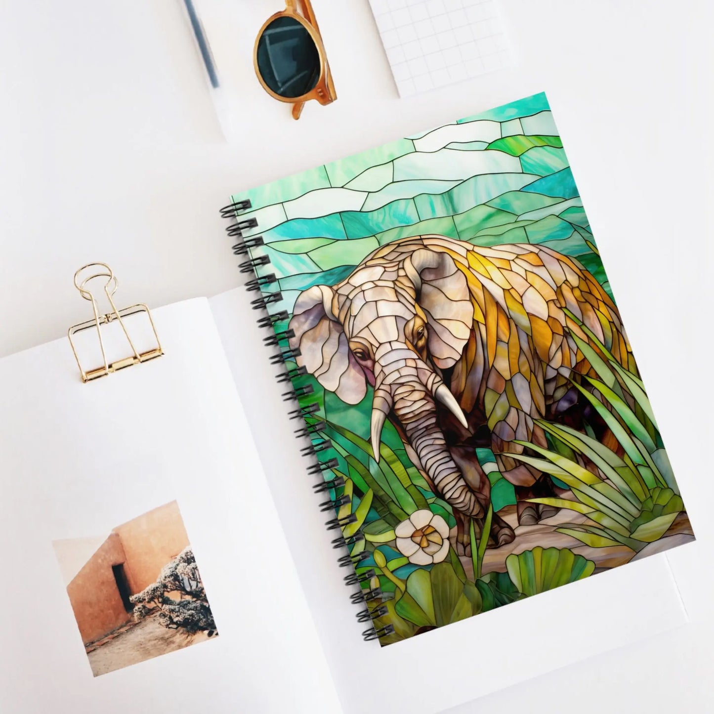 The Stained Glass Elephant Spiral Notebook, featuring a vibrant design with greenery, makes an ideal gift. Its beautifully displayed on a white surface with a photo, clip, sunglasses, and small notepad.