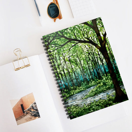 A flat lay features the Stained Glass Forest Spiral Notebook with a mosaic forest cover, paired with sunglasses, a grid-patterned notepad, and an open book displaying an image of an orange building.