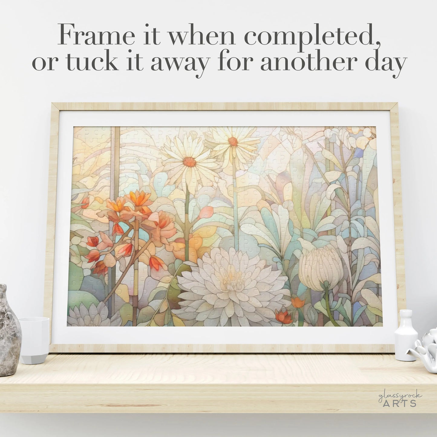 The Spring Botanicals Jigsaw Puzzle showcases vibrant, pastel-hued flowers like daisies and chrysanthemums, resembling a stained glass design. Displayed in a frame on a light wooden shelf with small decor, it features the text: Frame it when completed or tuck it away for another day.