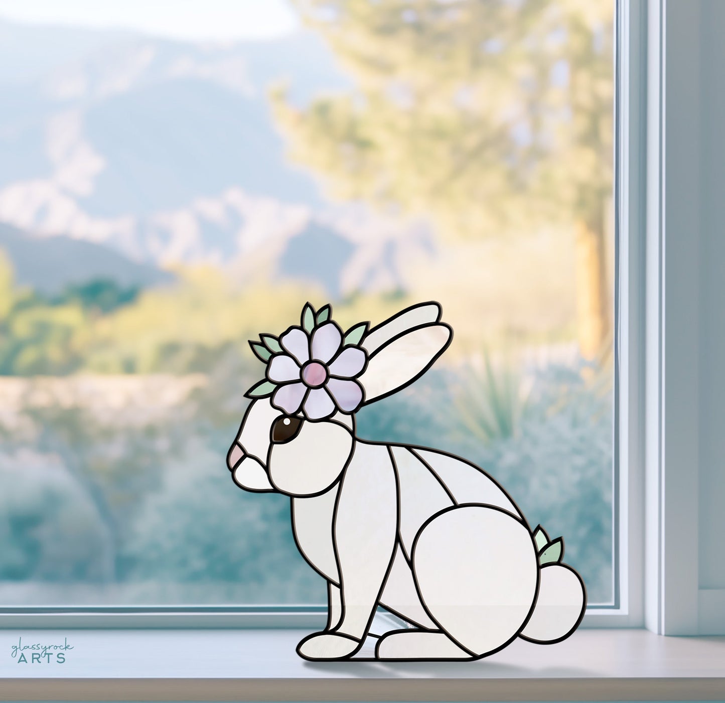 The Spring Rabbit Bunny Stained Glass Rabbit Pattern decorates a window ledge with its flower-crowned spring rabbit. An out-of-focus scene of trees and distant mountains adds tranquility. Its the perfect DIY Easter gift to brighten any room.