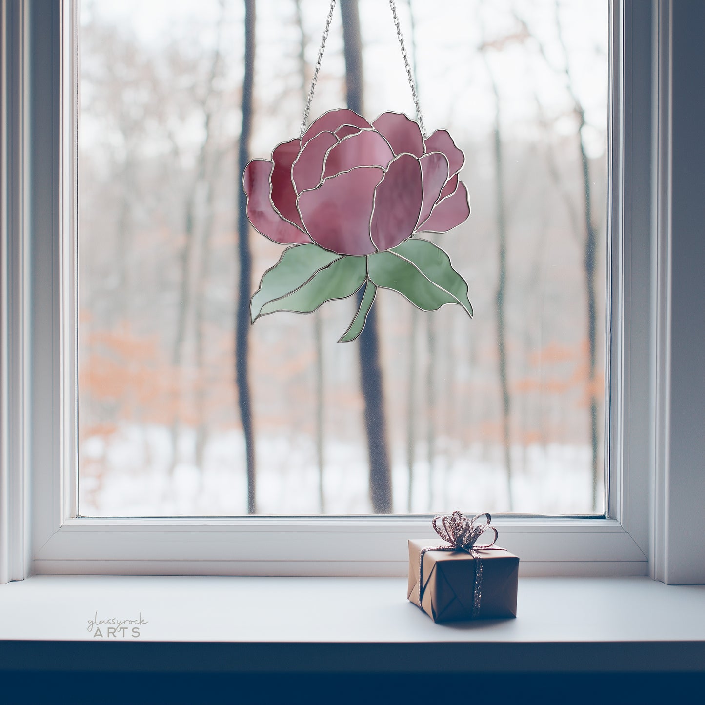 A Summer Rose Stained Glass Pattern sun catcher hangs in the window against bare trees, while a gift box with decorative ribbon sits on the windowsill. This serene scene invites thoughts of crafting with stained glass templates.