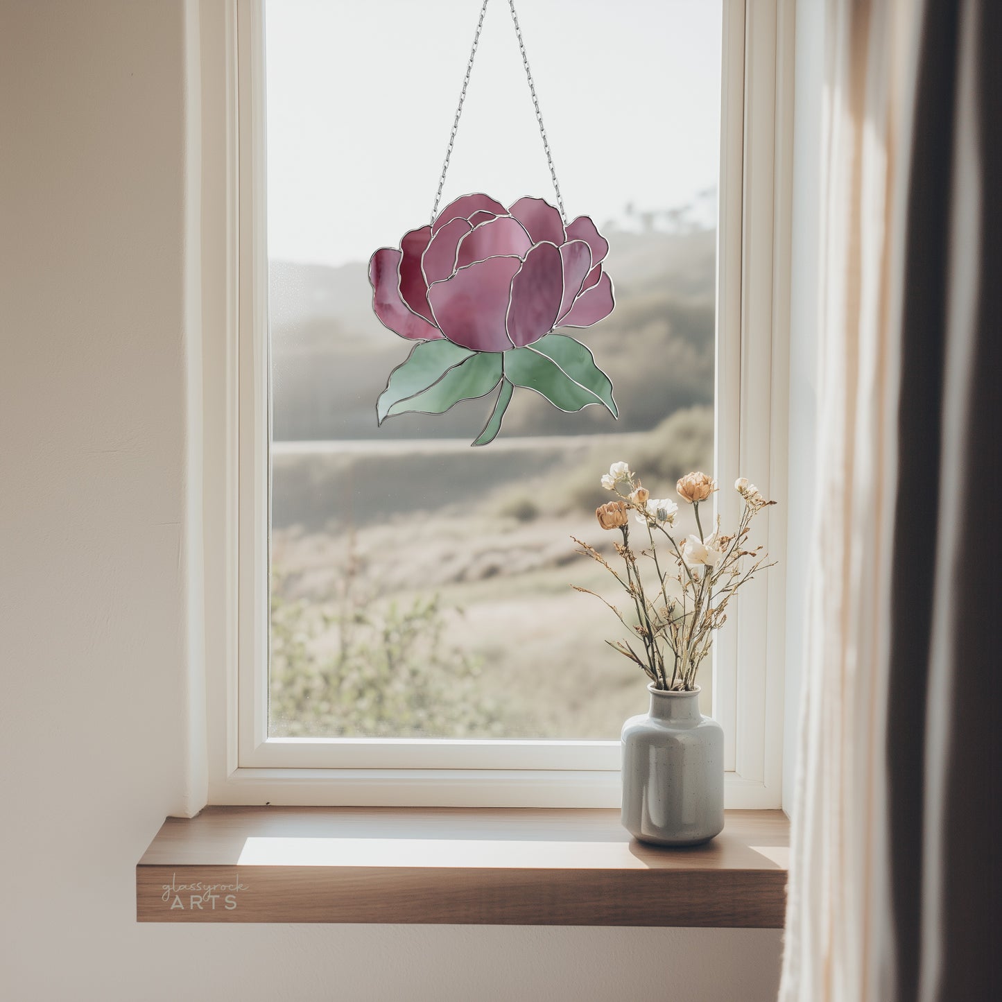 A window featuring the Summer Rose Stained Glass Pattern elegantly hangs at the top, with a blurred landscape outside. A small vase with dried flowers rests on the windowsill, framed by soft curtains, enhancing this charming view.