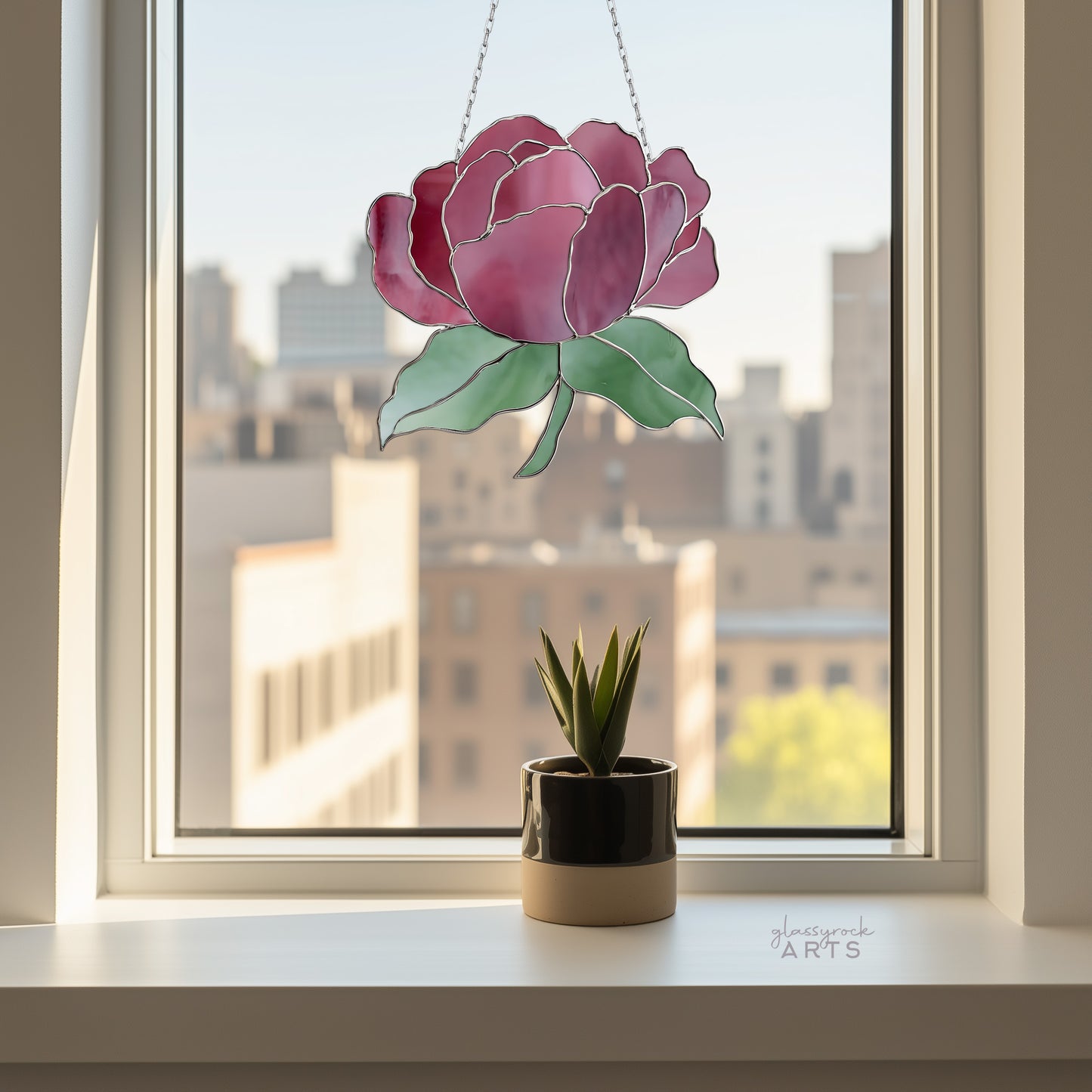 A potted succulent rests on a windowsill beside a Summer Rose Stained Glass Pattern sun catcher, with the cityscape softly blurred in the background, creating a tranquil urban scene.