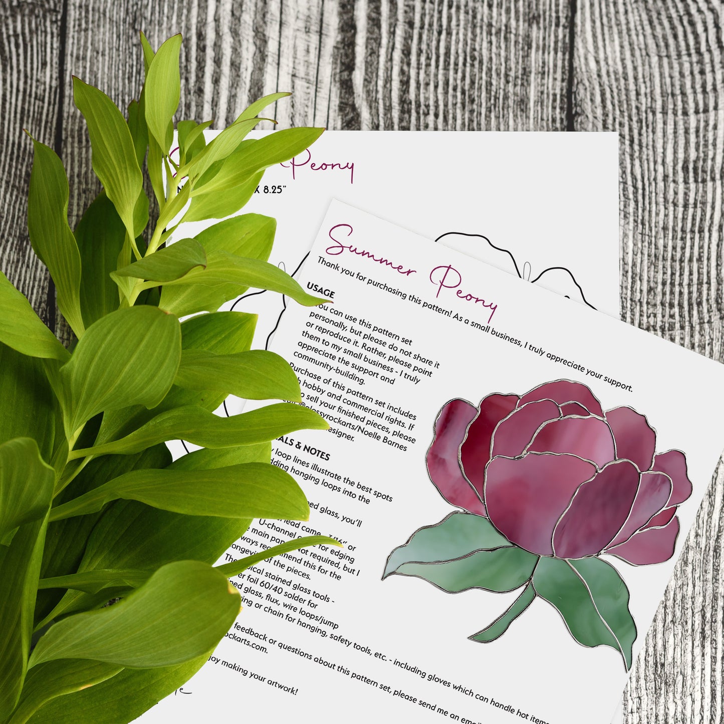 A leafy green plant is next to two papers on a wooden surface. The top paper, titled Summer Rose Stained Glass Pattern, features a vibrant illustration and details its usage, perfect for inspiring stained glass templates or printable patterns.