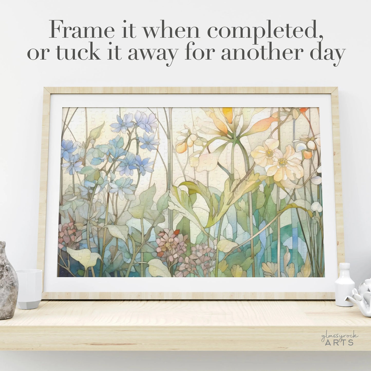 A Summer Wildflowers Jigsaw Puzzle framed artwork with blue, yellow, pink flowers, and green leaves is displayed on a shelf among decorative pieces. The text reads, Frame it when completed, or tuck it away for another day.