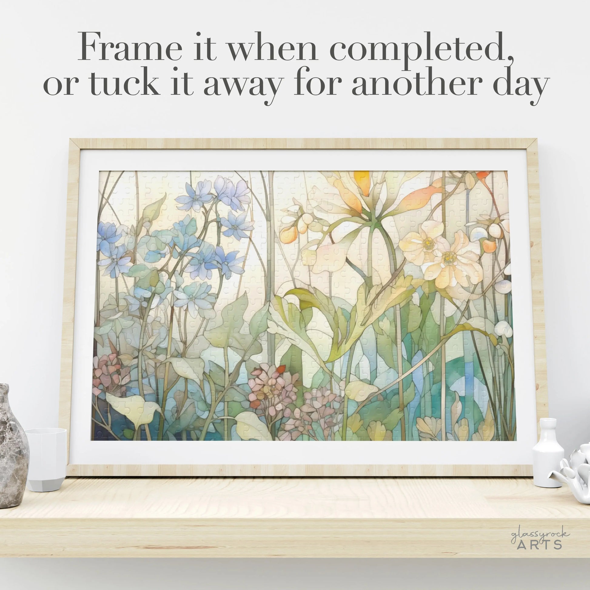A Summer Wildflowers Jigsaw Puzzle framed artwork with blue, yellow, pink flowers, and green leaves is displayed on a shelf among decorative pieces. The text reads, Frame it when completed, or tuck it away for another day.