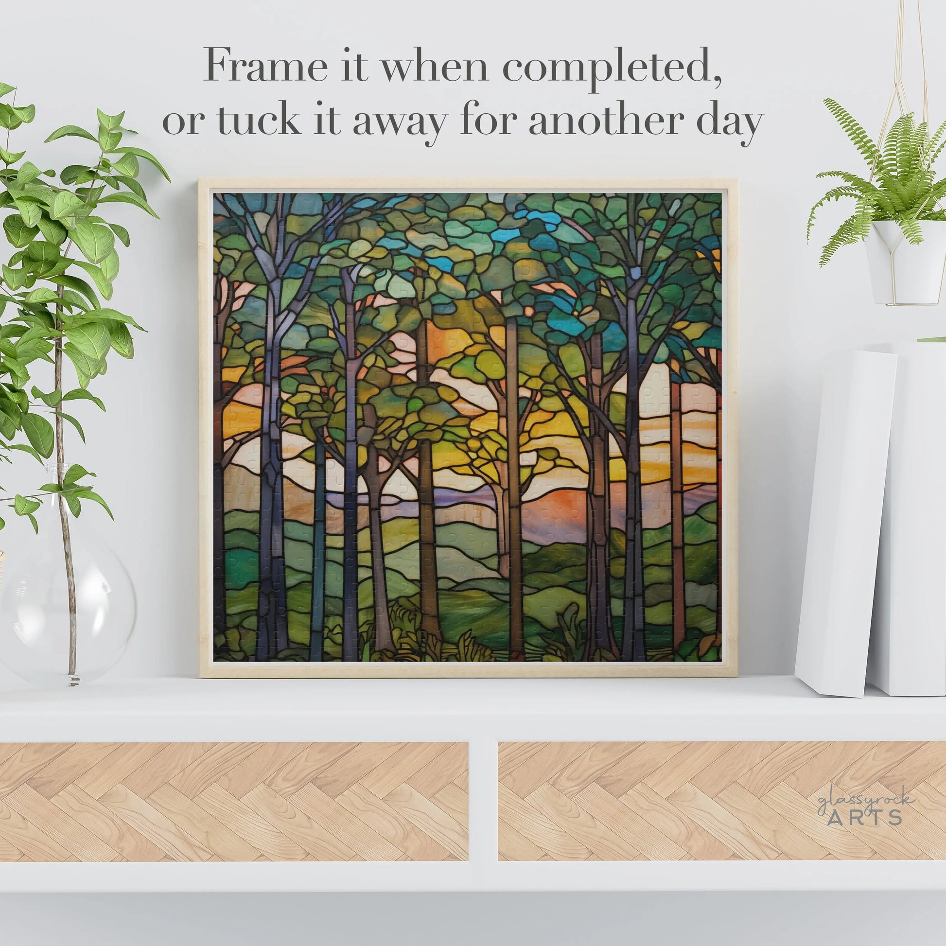 The Sunset Forest Jigsaw Puzzle, styled like stained glass art, rests elegantly on a white shelf. Its flanked by two potted plants and decorative accents. The text above suggests: Frame it when finished or store it for later—an ideal gift for nature enthusiasts.
