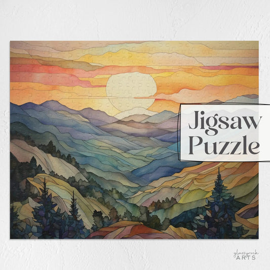 The Sunset Mountains Jigsaw Puzzle displays a vibrant landscape at dusk with orange, pink, and yellow skies over dark green pines. The Jigsaw Puzzle text appears on the right. Ideal for nature enthusiasts.