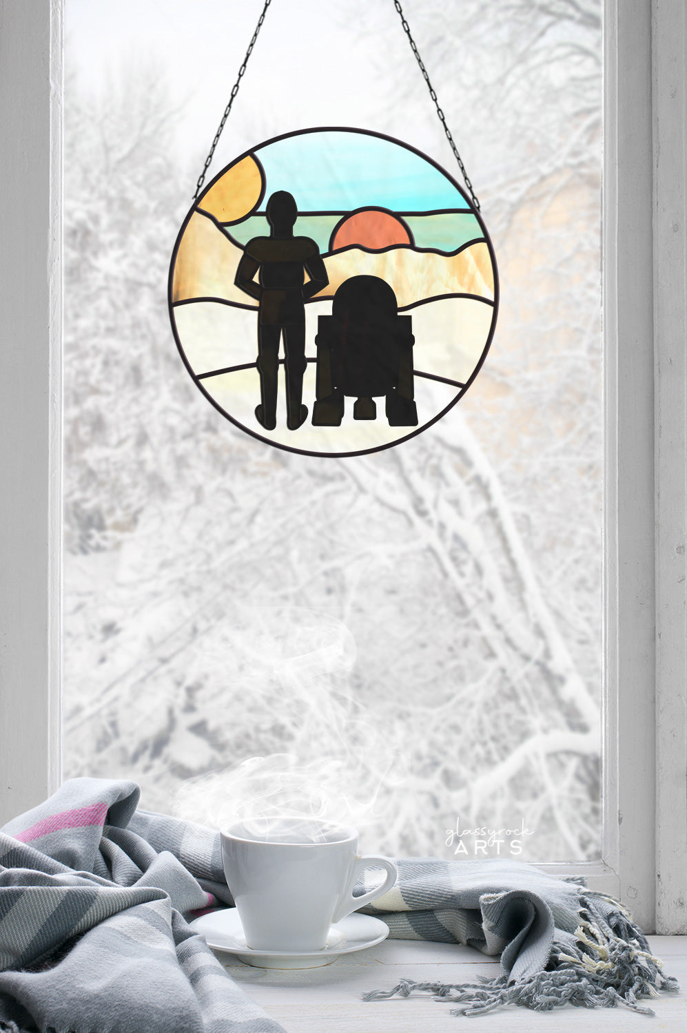 The Tatooine Landscape Stained Glass Pattern adorns a frosty window, highlighting silhouettes of two iconic sci-fi characters against a vibrant backdrop. Below, a steaming cup and soft blanket on the ledge create a cozy atmosphere.