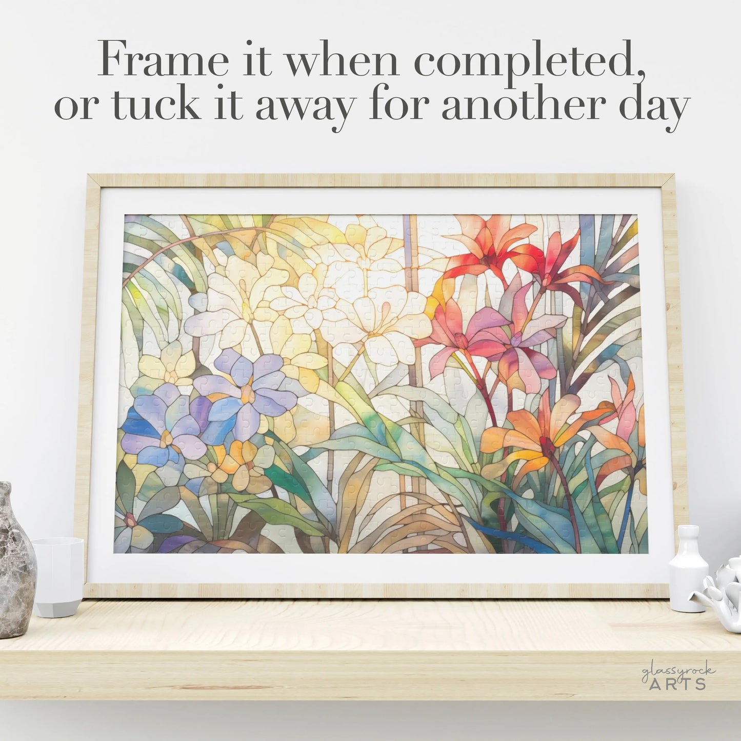 The Tropical Blooms Jigsaw Puzzle, showcasing vibrant stained glass-style flowers in purple, white, and red with green leaves, sits on a light wooden shelf. Above it reads, Frame it when completed or tuck it away for another day. A perfect art gift for any occasion.