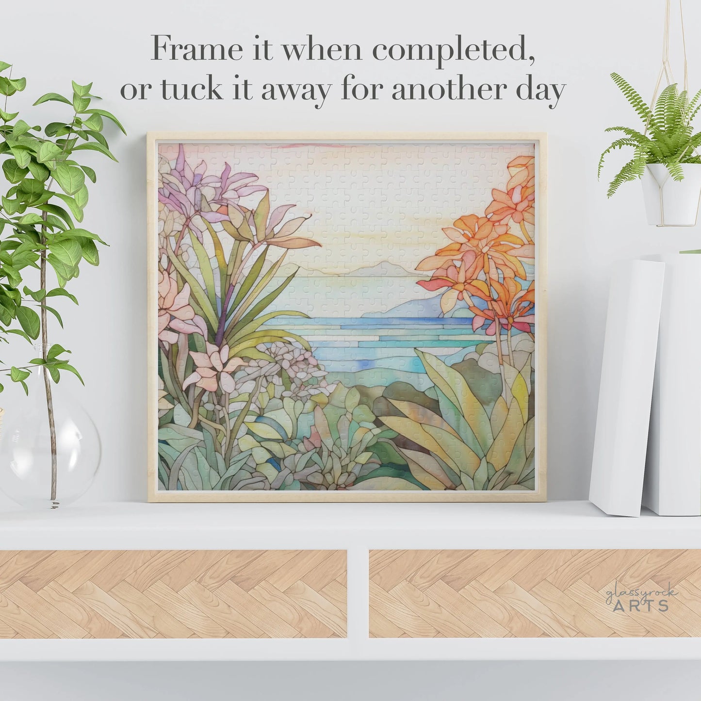 The Tropical Botanicals Jigsaw Puzzle, featuring a stained glass-inspired landscape with watercolor tropical plants, greenery, and a coastal view, is displayed on the shelf. The text suggests: Frame it when completed or tuck it away for another day.