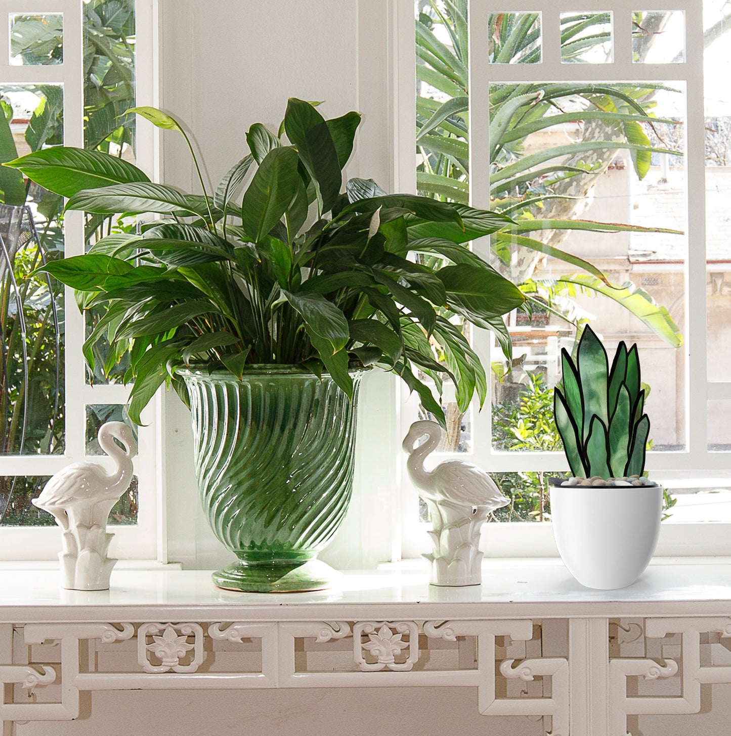 A decorative windowsill displays a large green 3D Snake Plant Leaves Stained Glass Pattern in a textured vase, flanked by two white swan figurines. A smaller plant in a matching white pot joins them, with lush greenery seen through the window as a backdrop.