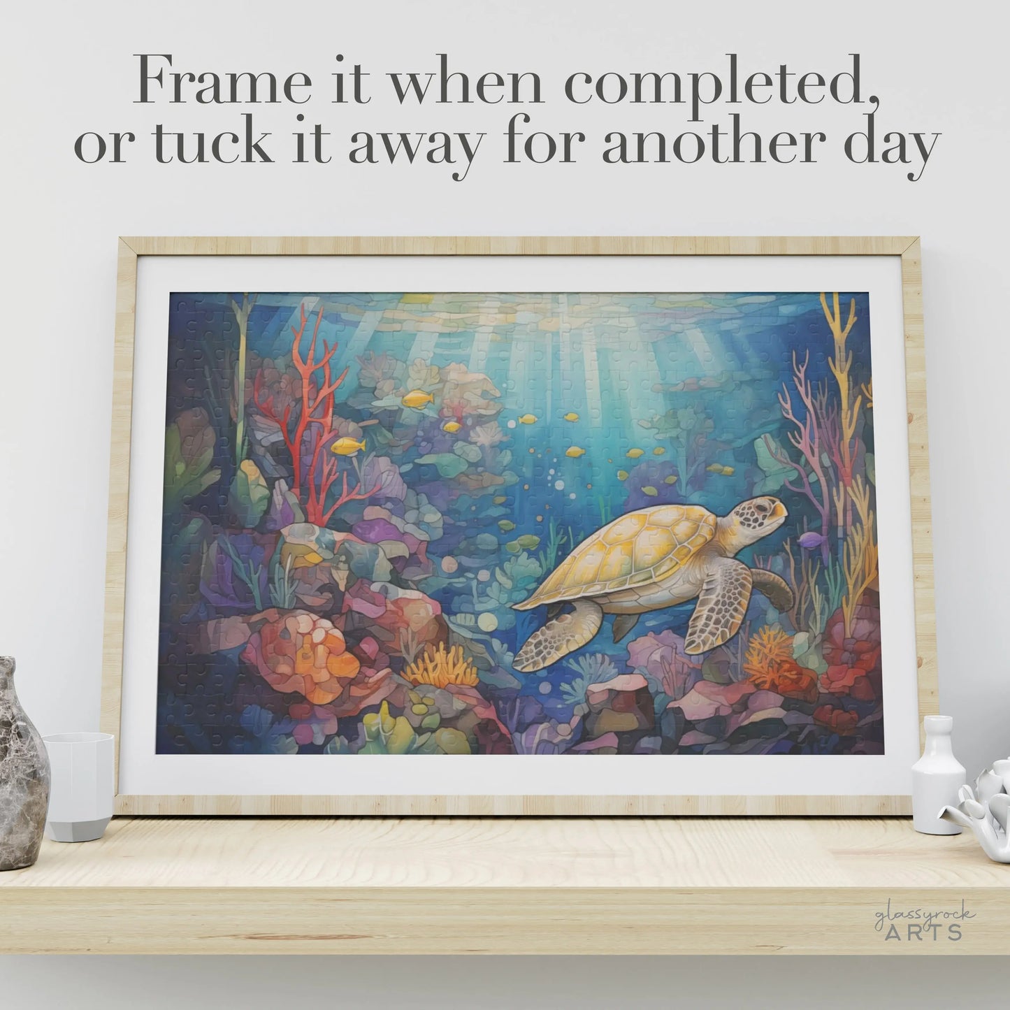 The Sea Turtle Reef Jigsaw Puzzle features a sea turtle swimming amidst colorful coral and fish. It comes with a light wood frame for display. The text reads, Frame it when completed, or tuck it away for another day. Perfect for puzzle lovers, with a paintbrush and a small white jar nearby.