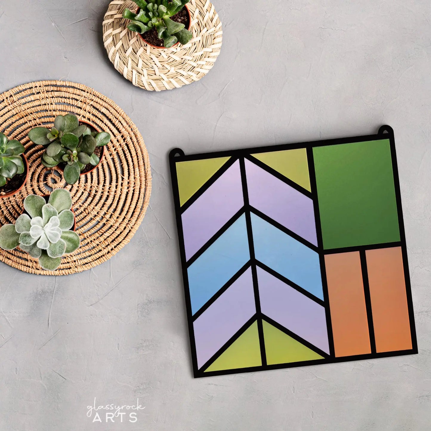 The display features a Stained Glass Square Suncatcher, showcasing geometric shapes in green, orange, blue, and purple on a gray surface. This Glowforge project, made with a laser cut file, is complemented by two baskets of succulents nearby.