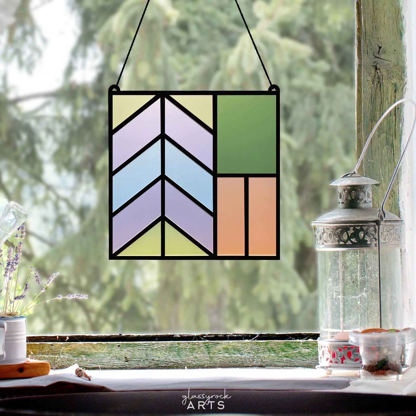 A Stained Glass Square Suncatcher SVG hangs in the window, flaunting a geometric pattern with pastel blue, purple, green, and peach tones. Beside it sits a decorative metal lantern on the sill, set against a blurred tree view—a perfect scene for your next Glowforge project.