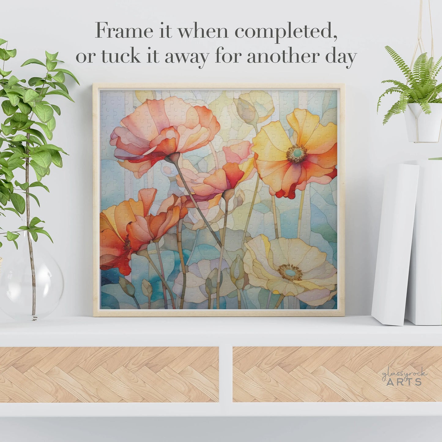 A Watercolor Poppies Jigsaw Puzzle featuring vibrant red, orange, and yellow petals beautifully graces a white shelf with a plant and decorative glass orb. The text above suggests to Frame it when completed, or tuck it away for another day. An ideal art gift for puzzle lovers.