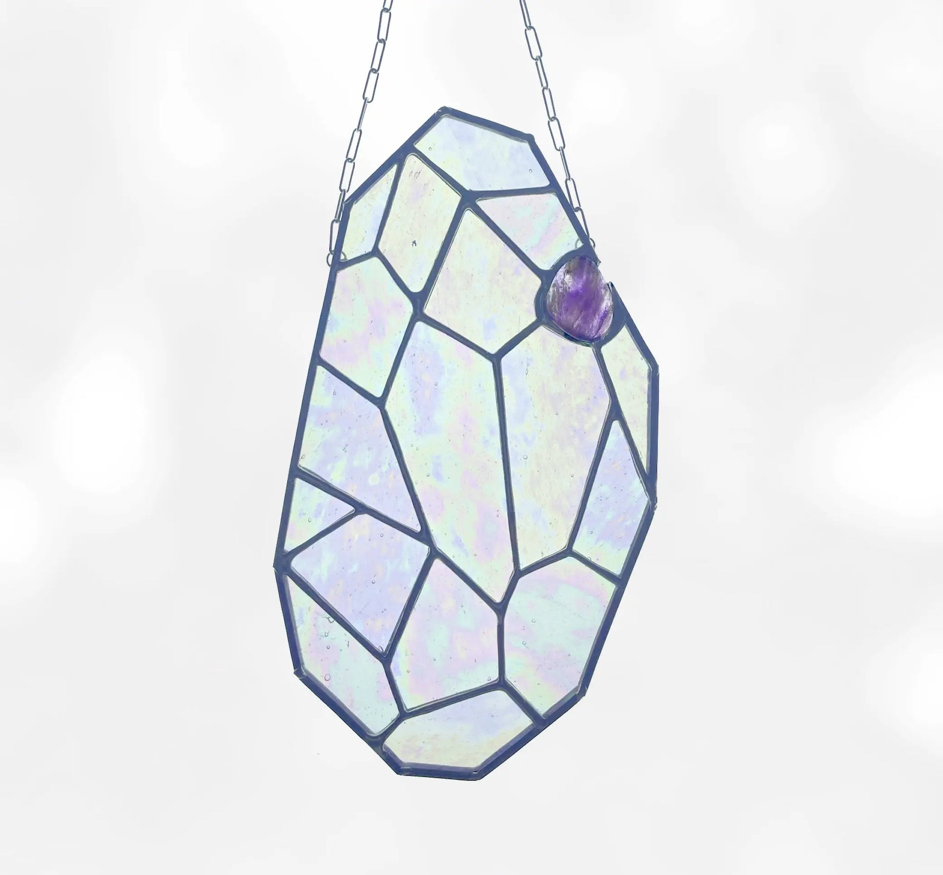 The Free Giant Gem, crafted by Noelle Barnes of GlassyRock Arts, is a decorative stained glass piece with an iridescent geometric design. It hangs by a chain and includes a small purple accent at the top against a soft, blurred white background.