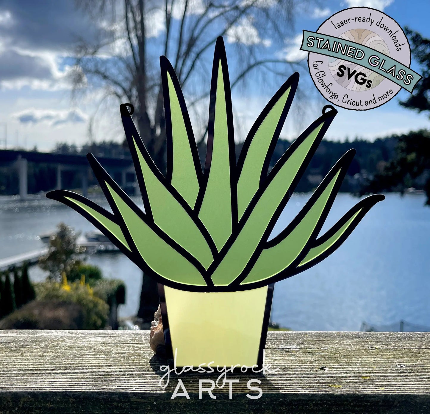 A vibrant Stained Glass Aloe Plant art piece, shaped like a potted succulent, rests on a wooden ledge overlooking a scenic body of water with a bridge and trees. A badge displays Stained Glass SVGs for crafting tools.