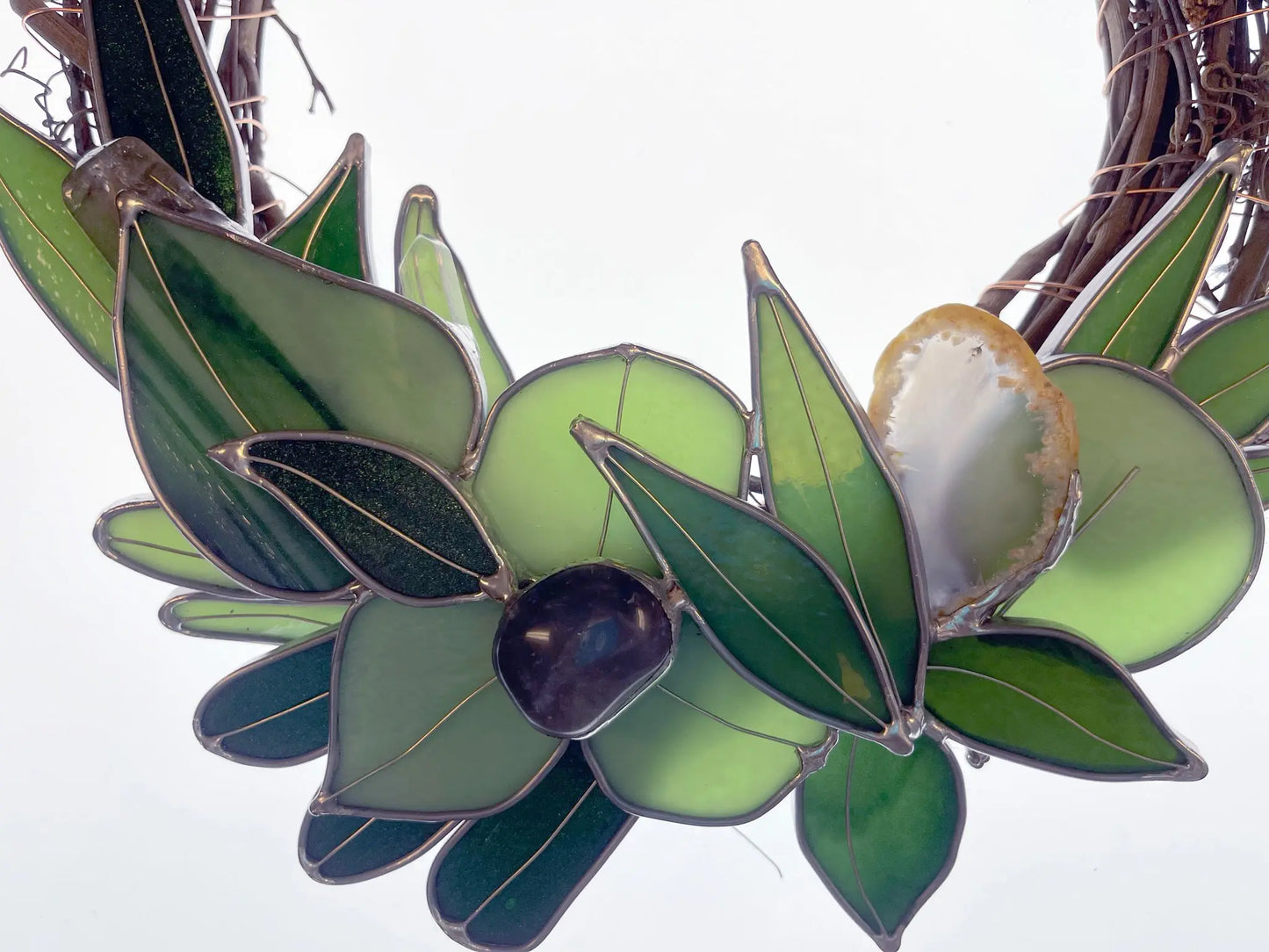 The Handmade Botanical Stained Glass Wreath features a close-up of leaf-patterned glass with a central black bead, accented by a brown and white shell on the right, set against a simple white background to enhance its artistic beauty.