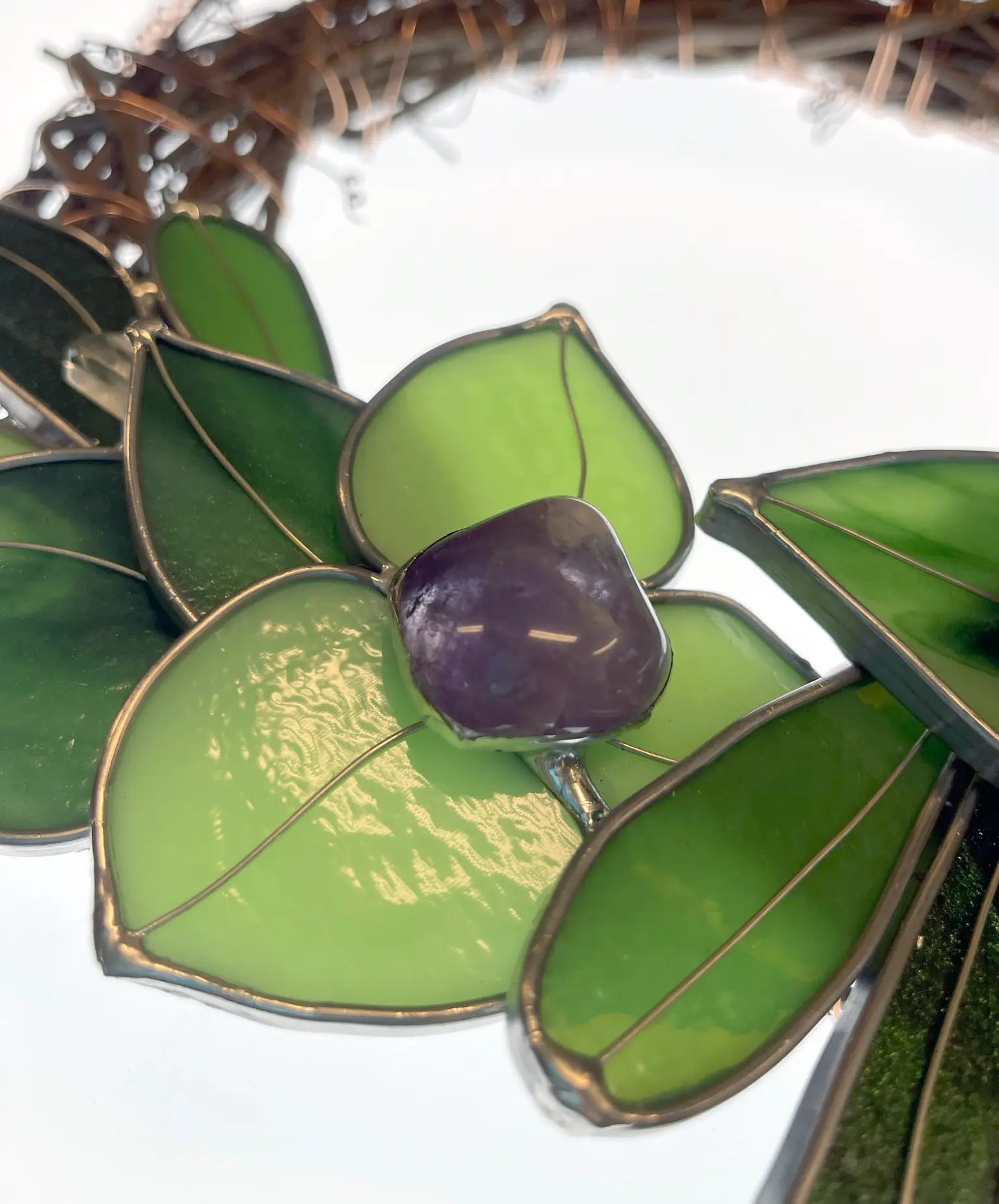 A close-up of the Handmade Botanical Stained Glass Wreath showcases a purple gemstone nestled among green, leaf-shaped pieces, all edged in thin metallic lines. The softly blurred background enhances the allure of this decorative treasure.