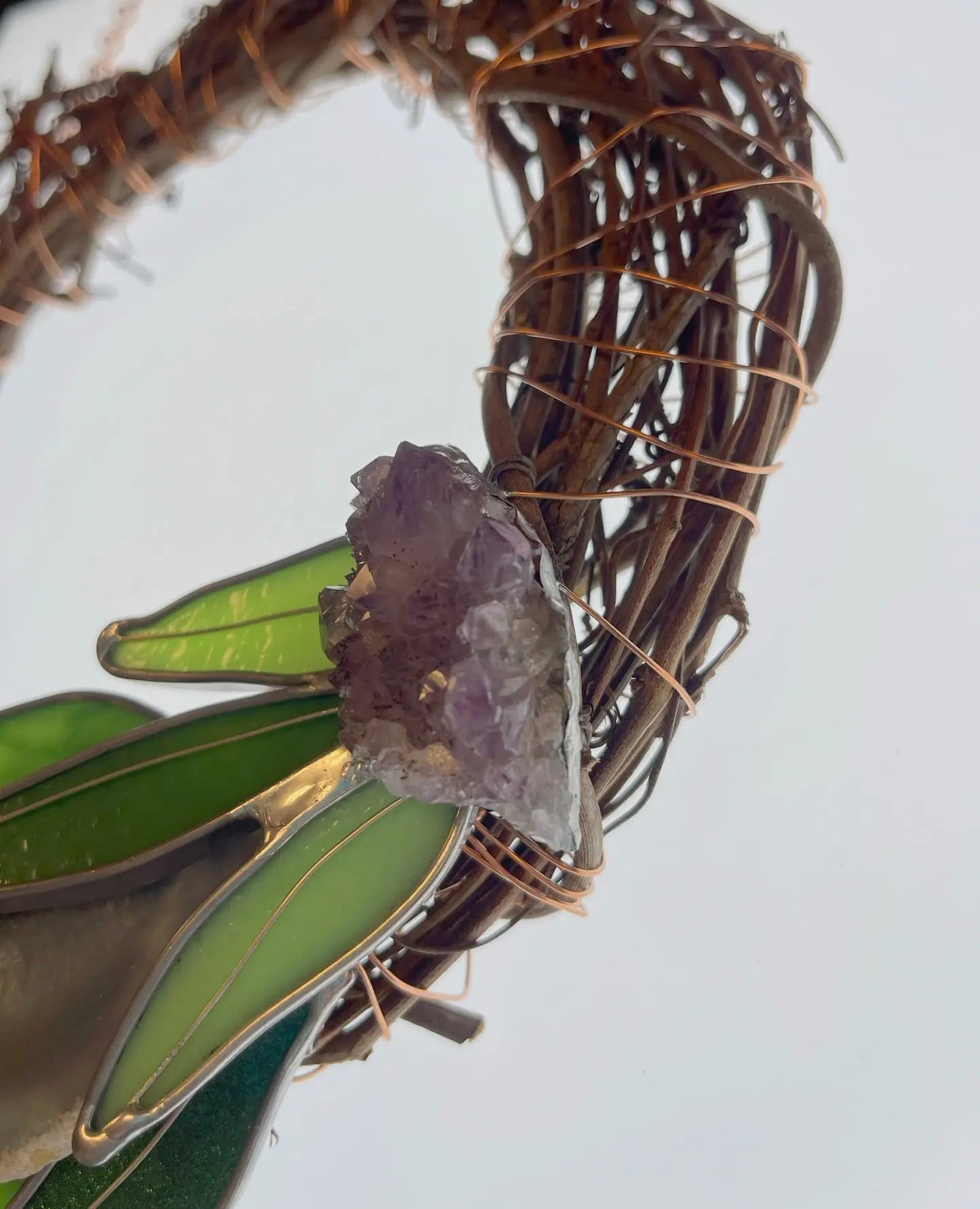 A close-up of the Handmade Botanical Stained Glass Wreath showcases clear, purple crystals on twisted wire and green-beige stained glass leaves, highlighting its rustic charm and botanical artistry.