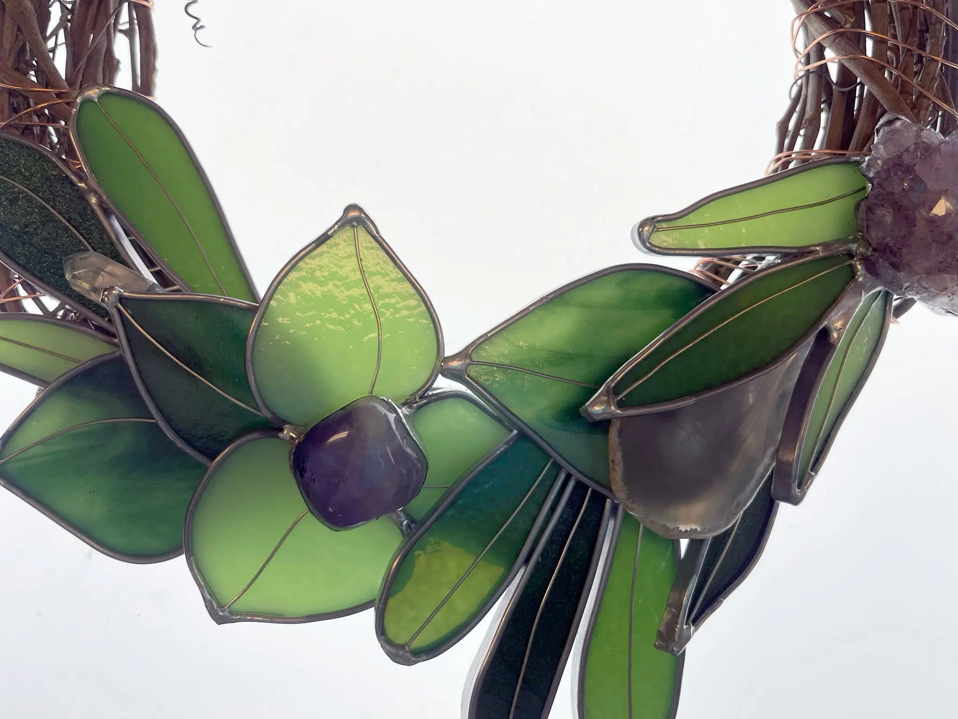 Close-up of the Handmade Botanical Stained Glass Wreath, showcasing green and light yellow leaves with a purple centerpiece. Thin, winding metal wires frame the design, creating an artistic motif against a light gray background.