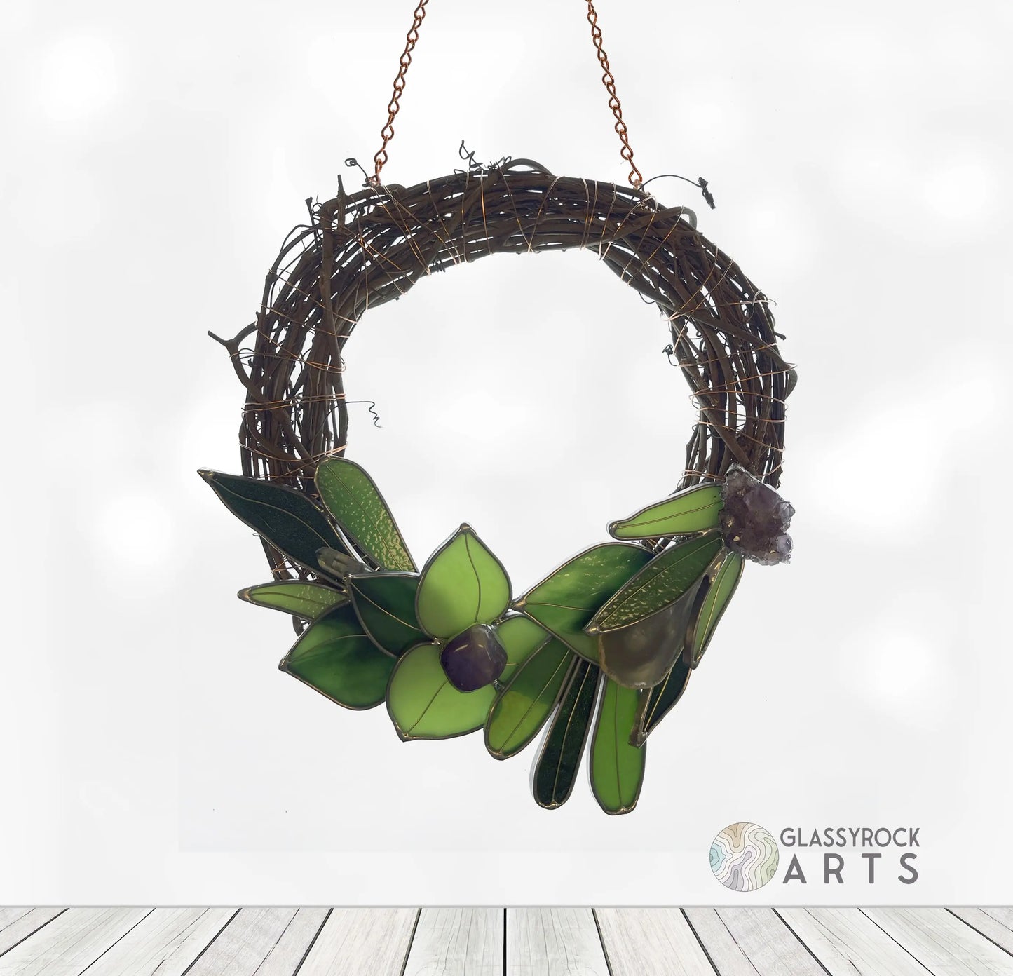 The Handmade Botanical Stained Glass Wreath features green leaves and a central flower, elegantly hanging from a chain. Crafted from twisted branches on a white background, it is adorned with the GLASSY ROCK ARTS logo in the bottom right corner.