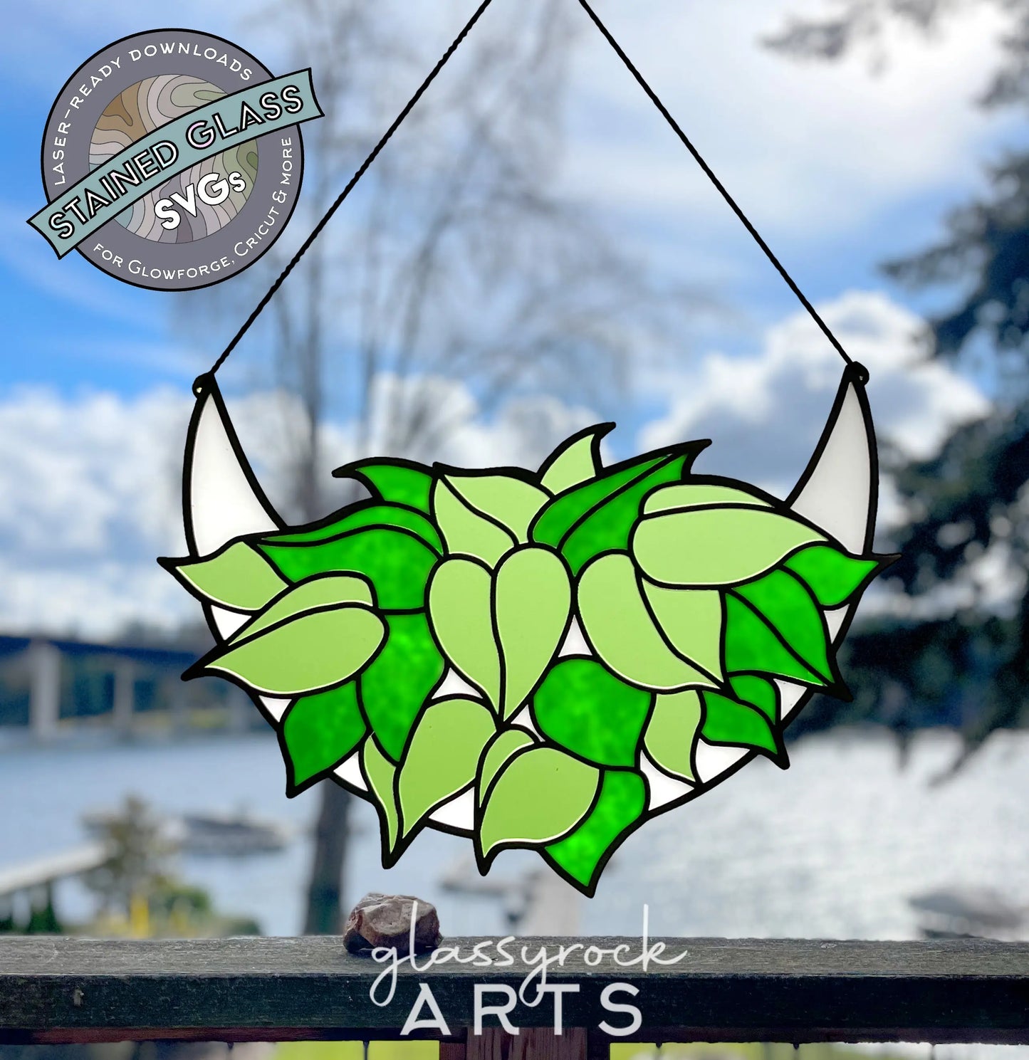 A stained glass design of green and light green leaves, recalling a philodendron SVG, hangs before a blurred outdoor scene with trees and water. The image features Stained Glass Philodendron Crescent Moon SVG Files by glassyrock ARTS.