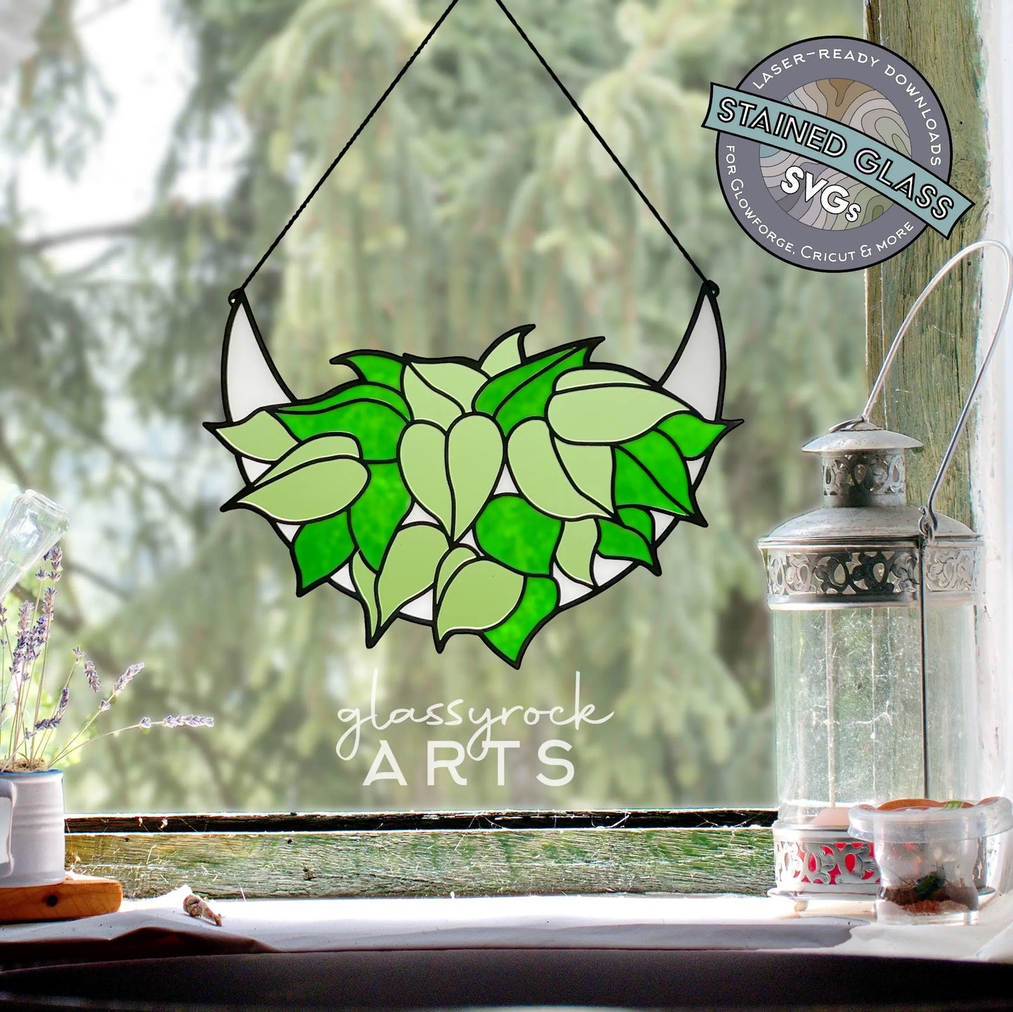 A Stained Glass Philodendron hangs by a window, complemented by a silver lantern and a small plant. The window view reveals lush greenery outside, while a circular logo at the top right highlights Stained Glass Philodendron Crescent Moon SVG Files.