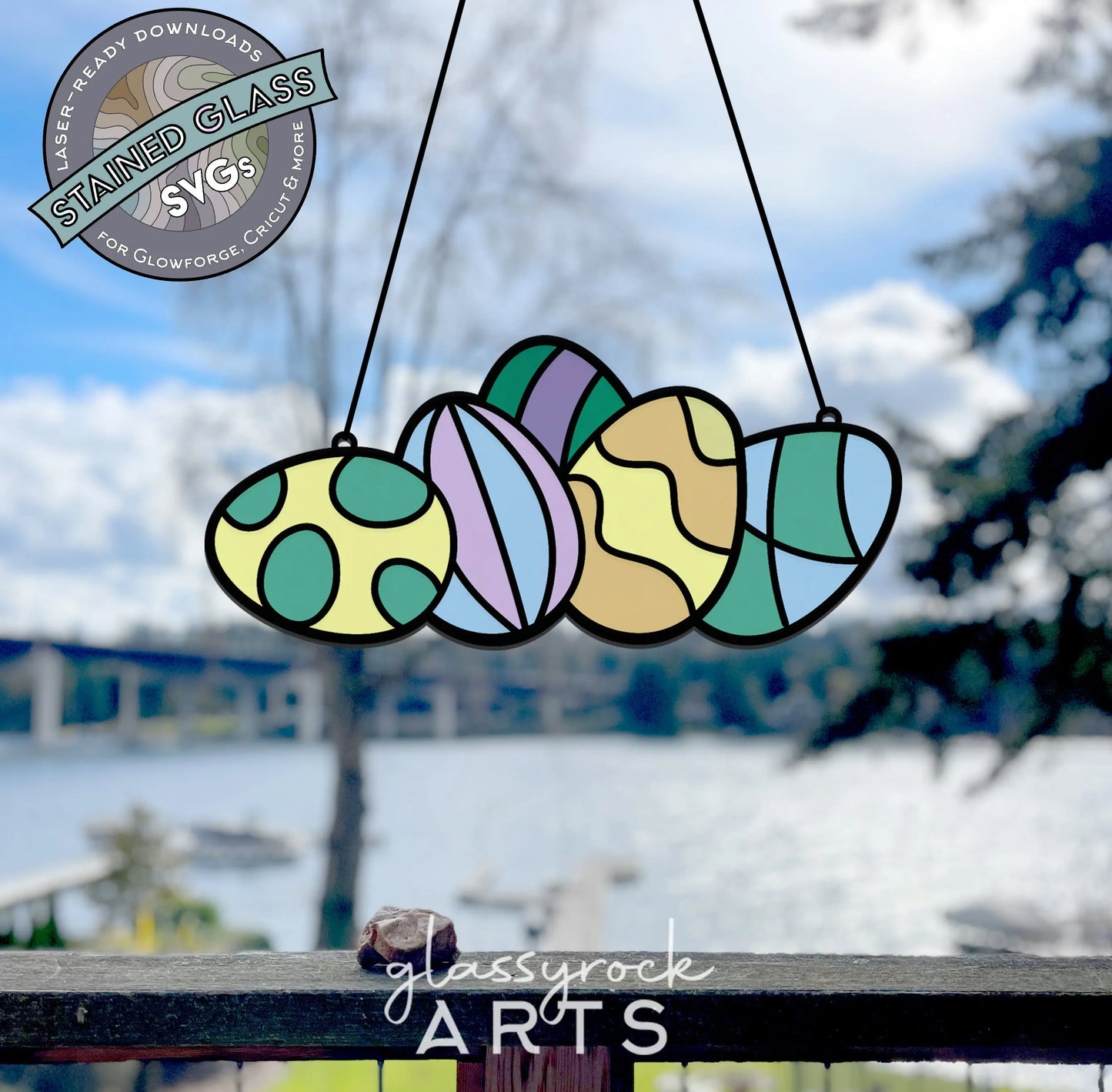 Vibrant stained glass Easter egg designs hang in front of a blurred outdoor scene with a lake, trees, and bridge. The text reads Stained Glass SVGs and glassyrock ARTS, making it perfect for a unique laser cut. Product: Stained Glass Easter Eggs SVGs.